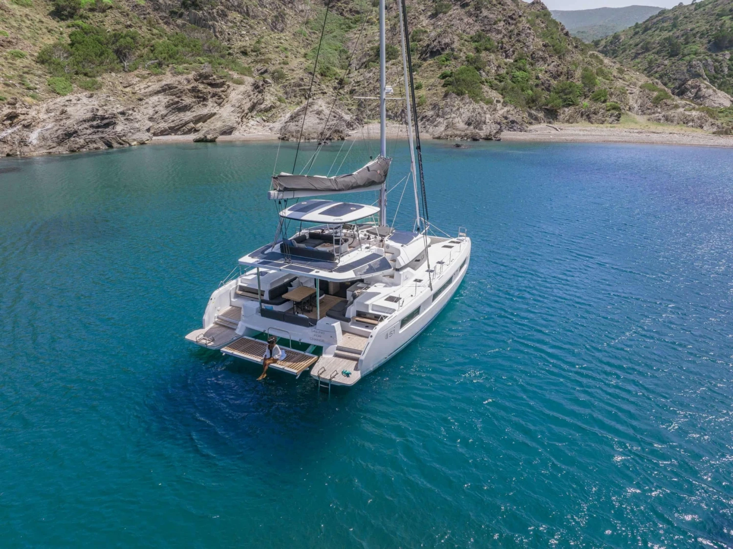 Yeşil tepelerle çevrili berrak turkuaz sularda, geniş güverteleri ve güneş panelleriyle dikkat çeken modern beyaz bir Lagoon 51 katamaran demirlemiş duruyor; kıç platformunda ayaklarını suya sokmuş bir kişi huzurlu anların tadını çıkarırken, bu tekne Singapur Yat Fuarı gibi prestijli etkinliklerin gözdesi olmaya aday.