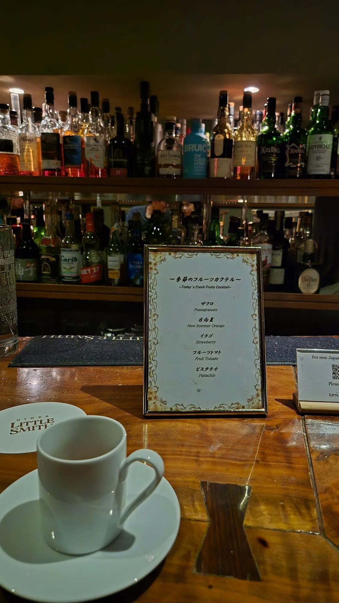 Loş ışıklı bir bar ortamında, ahşap tezgahın üzerinde duran beyaz bir fincan ve Ginza Little Smith logolu bardak altlığı, arka planda çeşitli içki şişeleriyle dolu rafların önünde, nar, yeni yaz portakalı, çilek, meyve domatesi ve fıstık gibi seçenekleri listeleyen Günün Taze Meyveli Kokteylleri menüsünü gösteren altın çerçeveli bir levhanın detaylı görüntüsü.