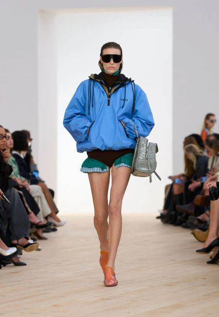 Podyumda yürüyen bir model, 2026 ilkbahar-yaz moda trendlerinden renk blokları akımını yansıtan, canlı mavi büyük beden bir anorak, kahverengi bir üst ve altından sarkan fırfırlı yeşil şortlar, geniş spor güneş gözlükleri, dokulu gri bir el çantası ve parlak turuncu ayakkabılarla güçlü bir siluet oluşturuyor.