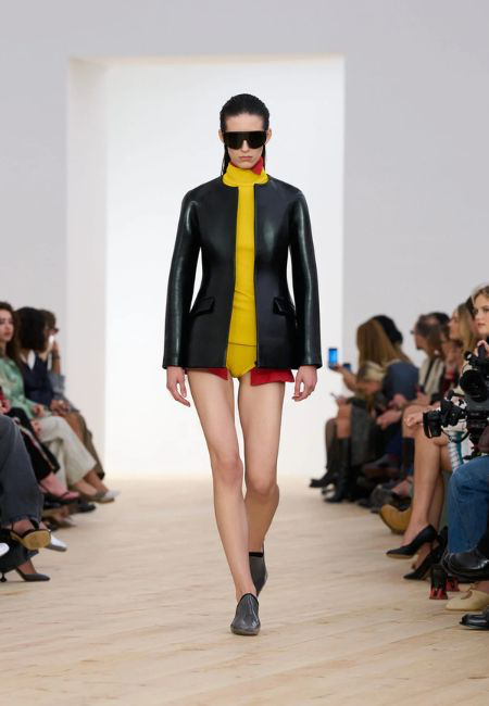 Loewe 2026 ilkbahar-yaz koleksiyonundan bir manken, podyumda yürürken, siyah deriden yapılmış minimalist ve keskin hatlara sahip bir ceket, altına giydiği canlı sarı yüksek yakalı bir elbise ve alt kısmından hafifçe görünen cesur kırmızı bir detayla renk blokları trendini modern bir şekilde sergiliyor.