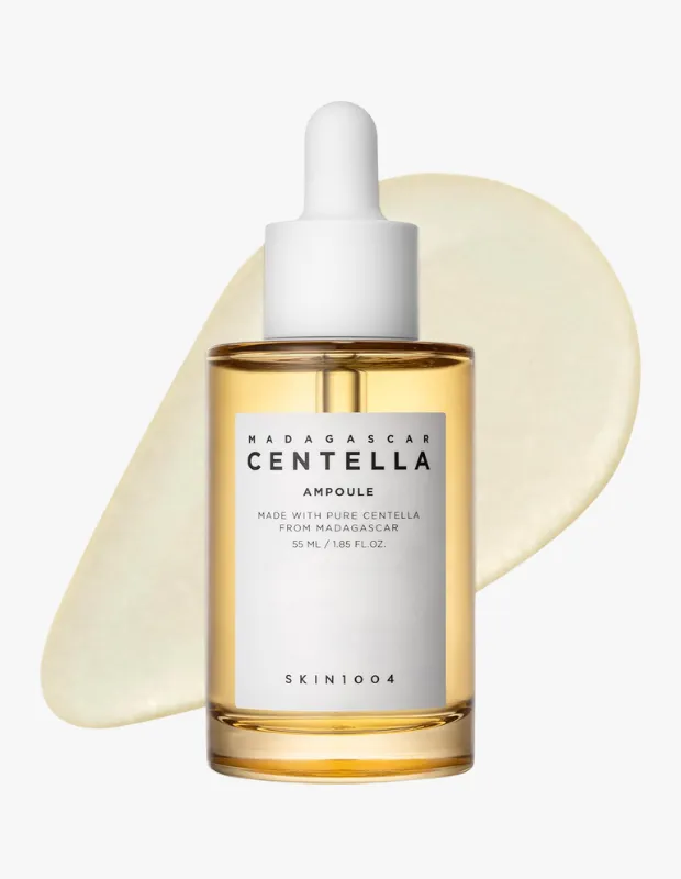 Beyaz bir zemin üzerinde, sarımsı kahverengi bir sıvı ile dolu, beyaz damlalıklı şeffaf cam bir şişe olan SKIN1004 Madagascar Centella Ampul, ürünün açık sarı, yoğun dokusunu sergileyen estetik bir damla iziyle zarifçe konumlandırılmıştır.