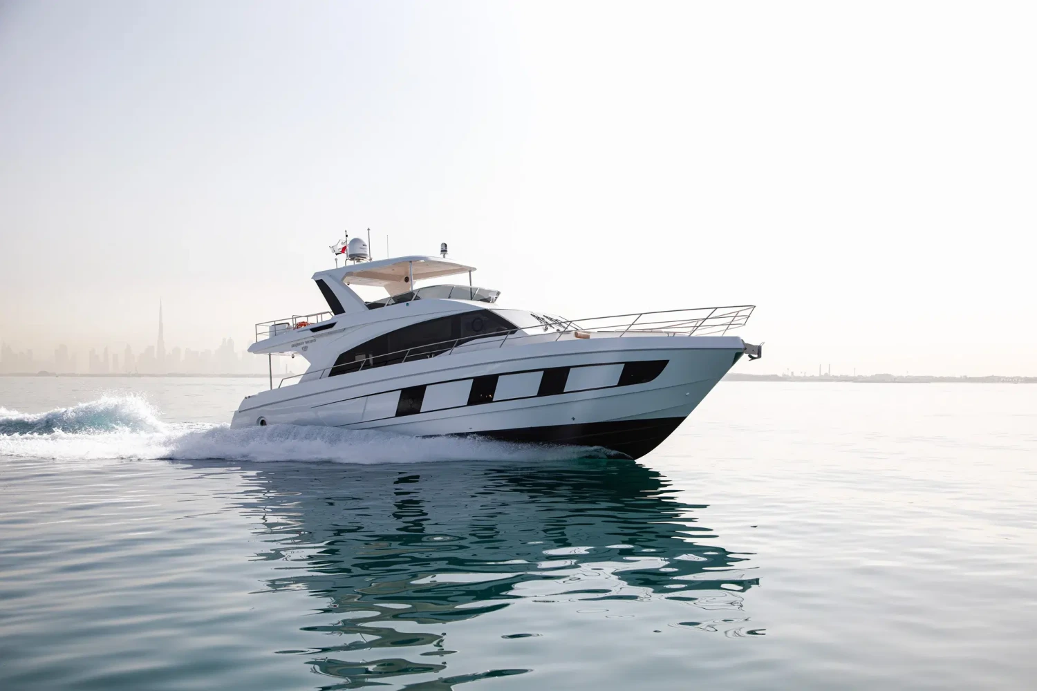 Beyaz Majesty 62 lüks motor yat, hafif dalgalı turkuaz denizde ilerlerken arkasında beyaz bir köpük izi bırakıyor; uzaklarda Dubai'nin puslu şehir silüeti seçiliyor ve bu görkemli deniz aracı Singapur Yat Fuarı bağlamında dikkat çekiyor.