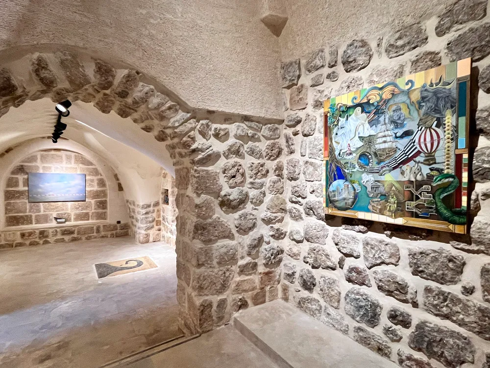 Mardin Bienali Eşik Sergisi'ne ev sahipliği yapan tarihi taş duvarlı bir galeride, sağ duvarda karmaşık, renkli ve sürreal bir tablo asılıyken, ilerideki kemerli niş içinde yatay bir ekran eseri sergilenmektedir.
