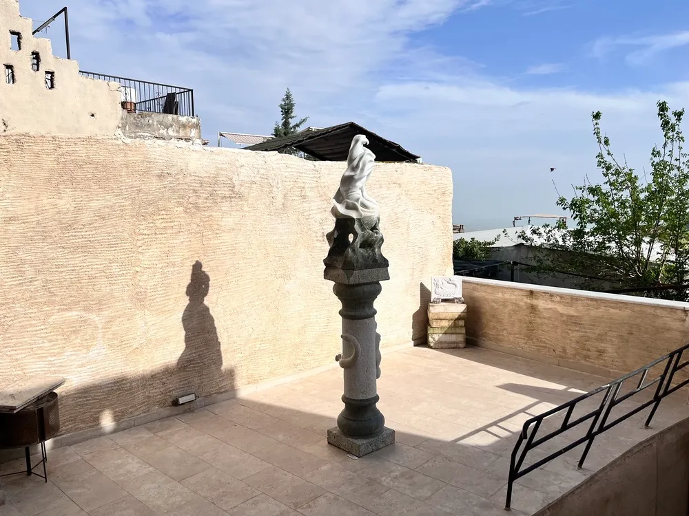 Mardin Bienali Eşik Sergisi kapsamında galerimizdeki güneşli bir terasta yer alan, dokulu bej bir duvarın ve taş karoların önünde, uzun bir kaide üzerindeki soyut beyaz heykelin ve heykelin solunda duvara vuran belirgin insan gölgesinin görüldüğü, Mardin'in genel dokusuna uygun bir açık hava düzenlemesi.