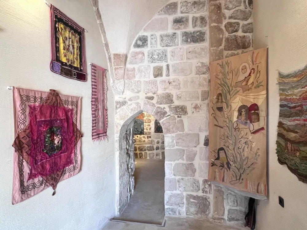 Mardin Bienali Eşik Sergisi'nden bir görünüm: Tarihi taş duvarlı bir galeride, geleneksel motiflerle modern yorumların harmanlandığı, pembe ve sarı tonlarında nakışlı kumaş panolar ile insan figürleri ve kuş işlemeli büyük bej bir goblenin sergilendiği bir koridor.