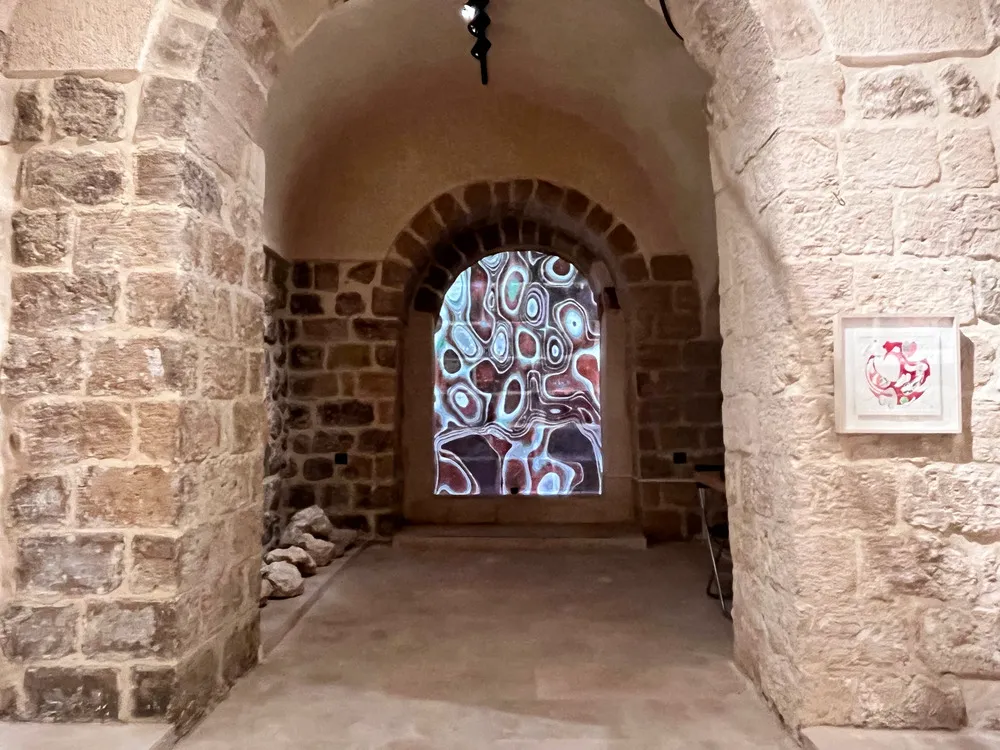 Taş duvarlı eski bir koridorda, kemerli bir geçidin içinde mavi, beyaz, kahverengi ve kırmızı tonlarda soyut, dalgalı desenler yansıtan bir dijital sanat enstalasyonu ve sağ duvarda asılı pembe tonlarda çerçeveli bir eser, Mardin Bienali'nin Eşik Sergisi'nden etkileyici bir görünüm sunuyor.