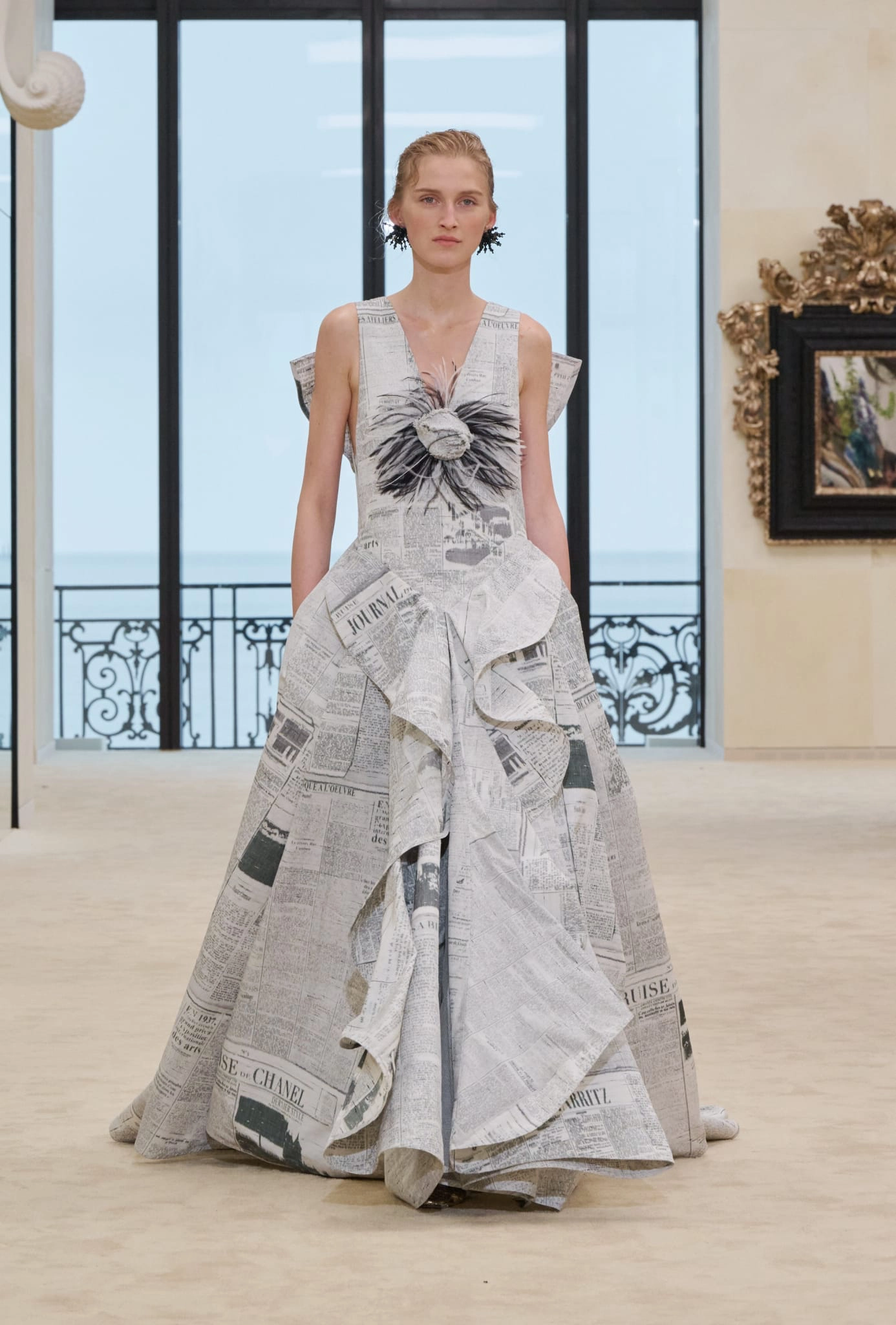 Matthieu Blazy'nin Chanel için tasarladığı ilk Cruise koleksiyonundan bir parça sergileyen genç model, Biarritz’deki defile alanında deniz manzaralı pencerenin önünde duruyor; modelin üzerindeki kabarık etekli, eski gazete baskılı, göğsü tüylü siyah-beyaz çiçek detaylı ve fırfırlı elbise, markanın tarihi köklerine dönüş temasını yansıtıyor.