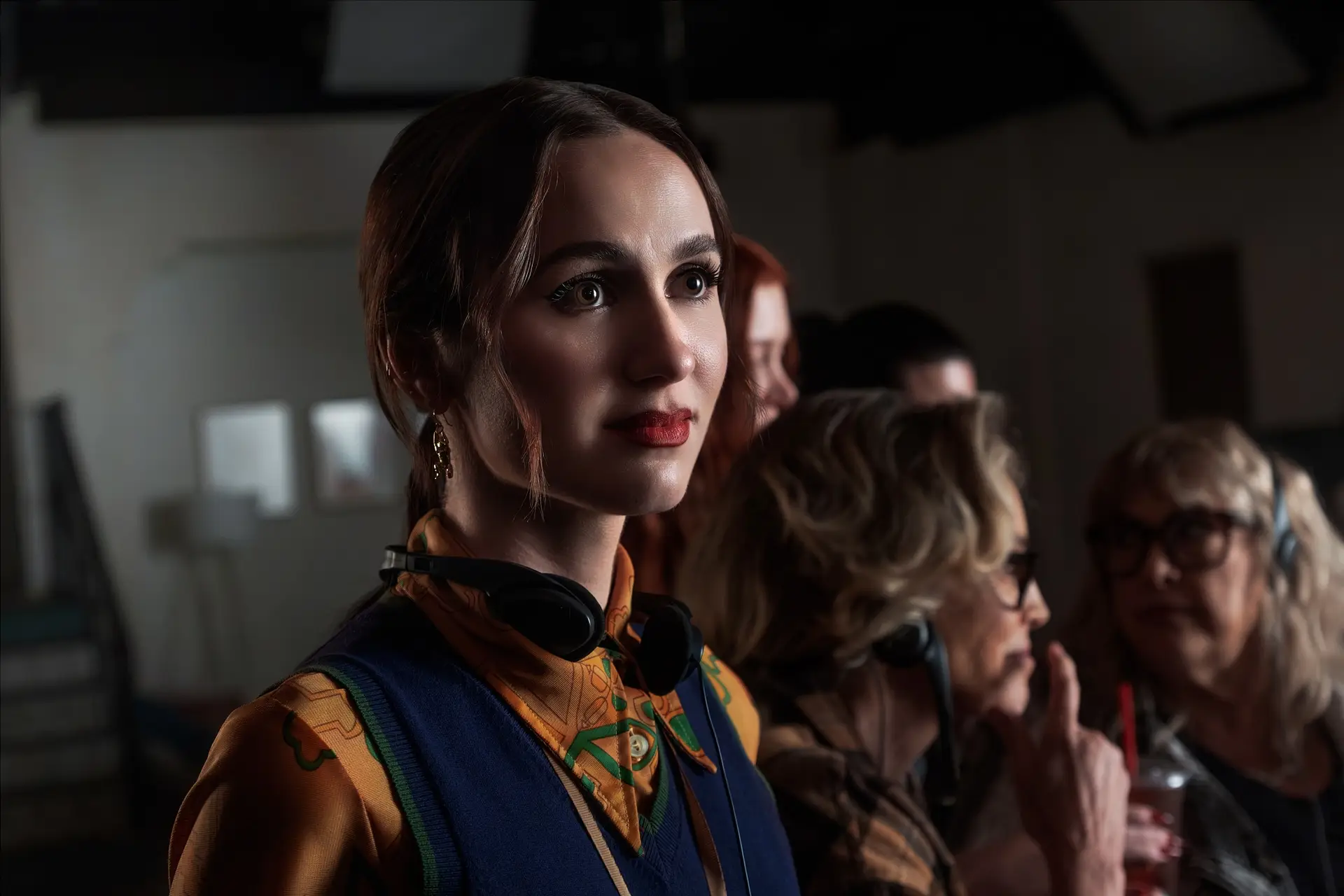 Euphoria 3. Sezon için bekleyiş devam ederken, Maude Apatow koyu kahverengi saçları, kırmızı ruju ve dikkatli bakışlarıyla öne çıkıyor; sarı desenli bir gömlek ve lacivert bir yelek giymiş, boynunda siyah kulaklıklar asılıyken, arkasında bulanıkça görünen diğer kişilerle birlikte bir ortamda.