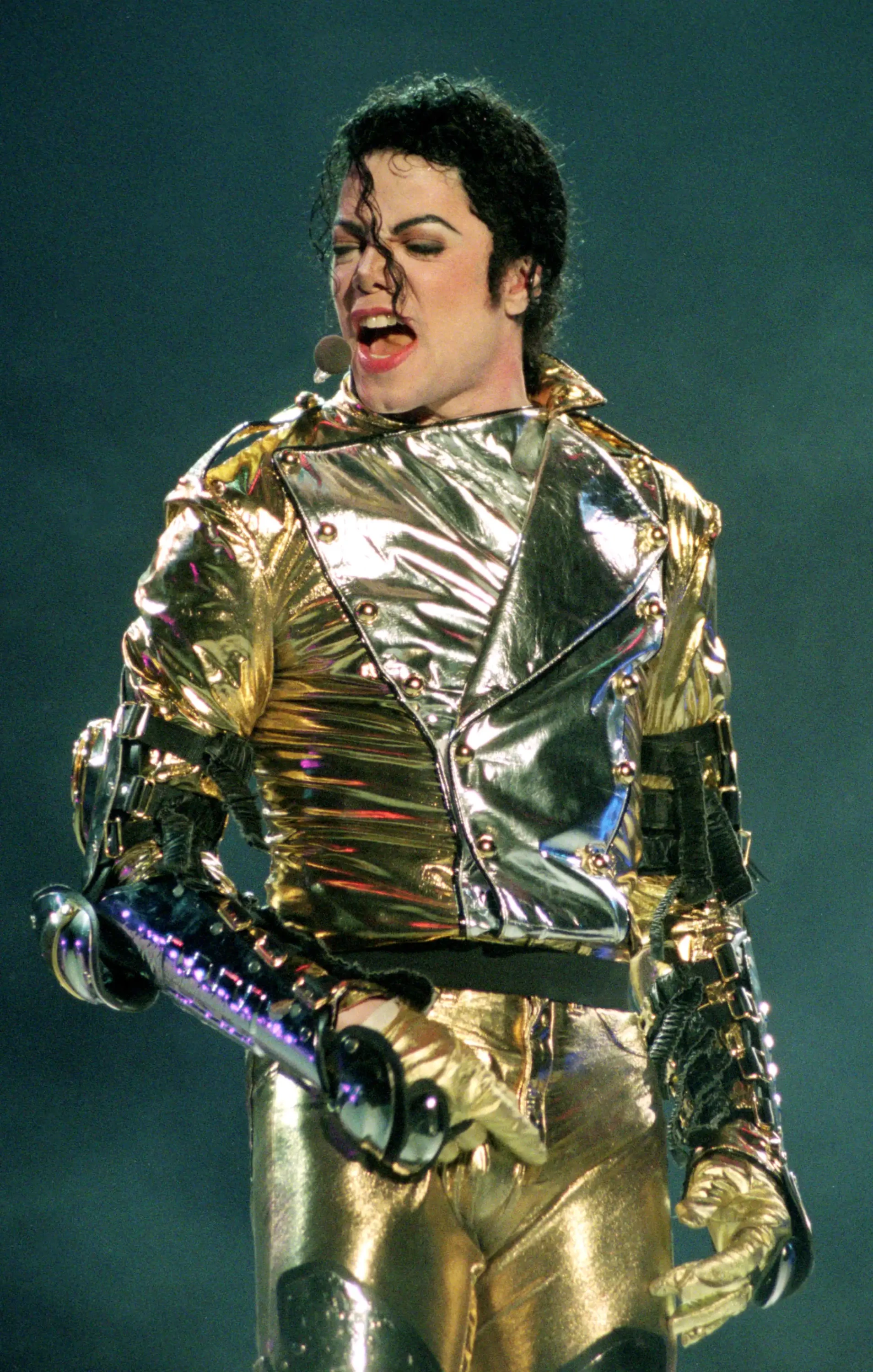 1993 Super Bowl XXVII sahnesinde, altın ve gümüş renkli metalik, zırh hissi veren görkemli kostümüyle, güçlü ve iddialı sahne stilinin zirvesinde şarkı söyleyen Michael Jackson'ın bu anı, onun ikonik tarzının ne kadar etkileyici olduğunu gösteriyor.