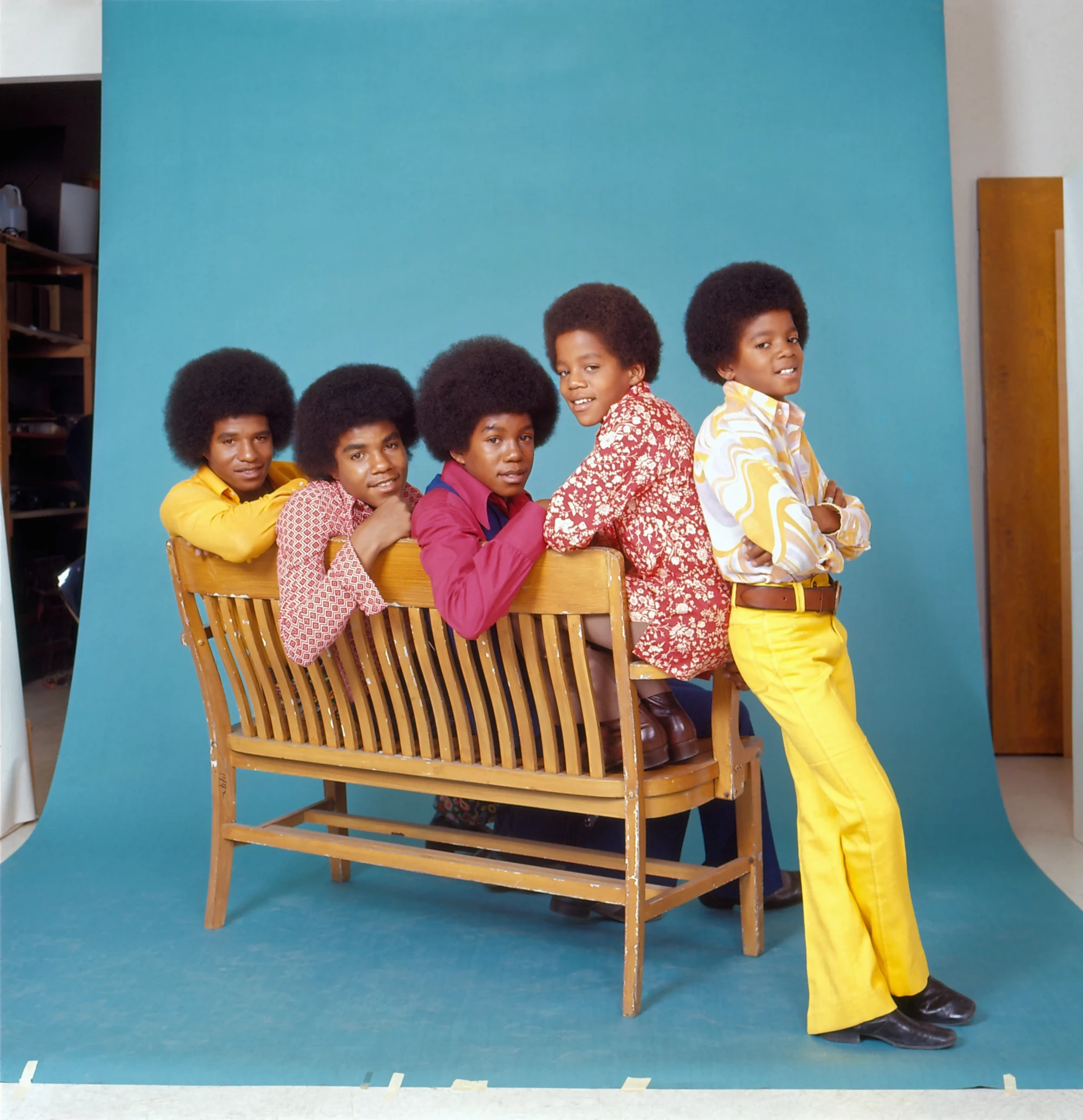 1972 civarında Los Angeles'ta çekilen bu stüdyo fotoğrafında, Jackson 5 üyeleri parlak mavi bir fon önünde ahşap banka yaslanmış, hepsi kabarık afro saçlara sahip. En sağda ayakta duran genç Michael Jackson, sarı desenli gömleği ve geniş paçalı pantolonuyla, grubun stil yolculuğunun başlangıcındaki canlı renkli ve uyumlu sahne tarzını sergiliyor.