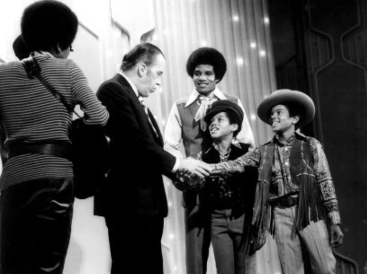 Siyah beyaz fotoğrafta, The Ed Sullivan Show sahnesinde genç Michael Jackson kovboy şapkası ve püsküllü yeleğiyle neşeyle sağa bakarken, programın sunucusu Ed Sullivan solunda onunla el sıkışıyor; arka planda Jackson 5 grubunun diğer üyeleri gururla gülümseyerek duruyor.