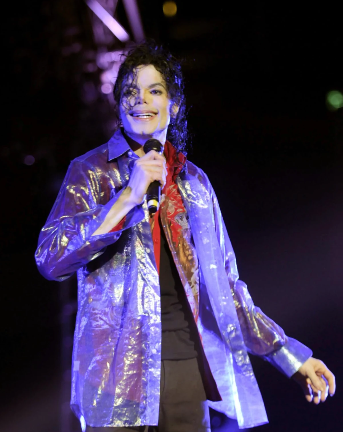 Michael Jackson, mor-mavi sahne ışıkları altında parlayan saydam bir gömlek giymiş, elinde mikrofonuyla geniş bir şekilde gülümserken, kıvırcık saçları yüzünü çerçeveliyor ve coşkulu bir konser performansı anında izleyicilerle göz teması kuruyor.