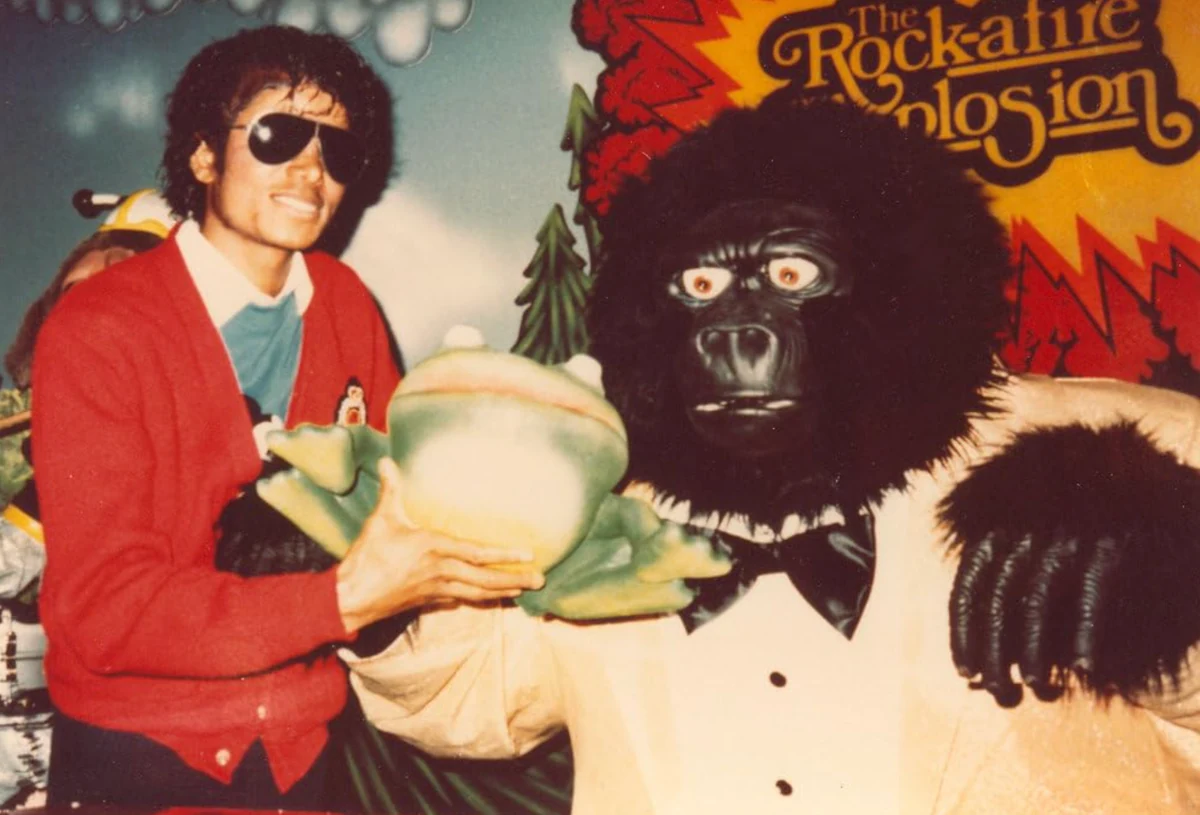 Yeşil bir kurbağa karakterini tutarken Rock-afire Explosion grubundan Fatz Geronimo animatronik gorilinin yanında parlakça gülümsüyor, arkada ise grubun logosu görülüyor.