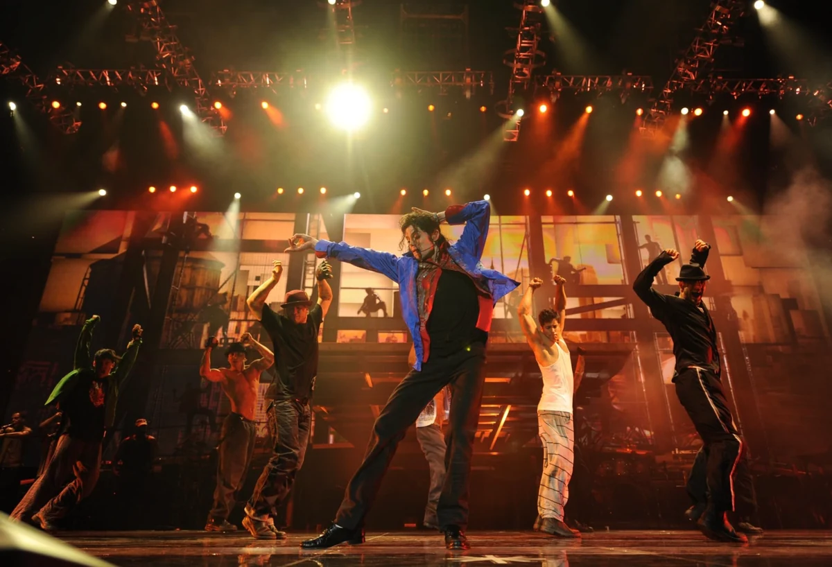 Michael Jackson, This Is It provalarından veya sahnesinden bilinen ikonik dans pozlarından birini sergilerken, parlak ışıklarla aydınlatılmış sahnede, arkasında kent manzarası görüntüsü ve etrafında dansçılarıyla coşkulu bir performans sergiliyor.