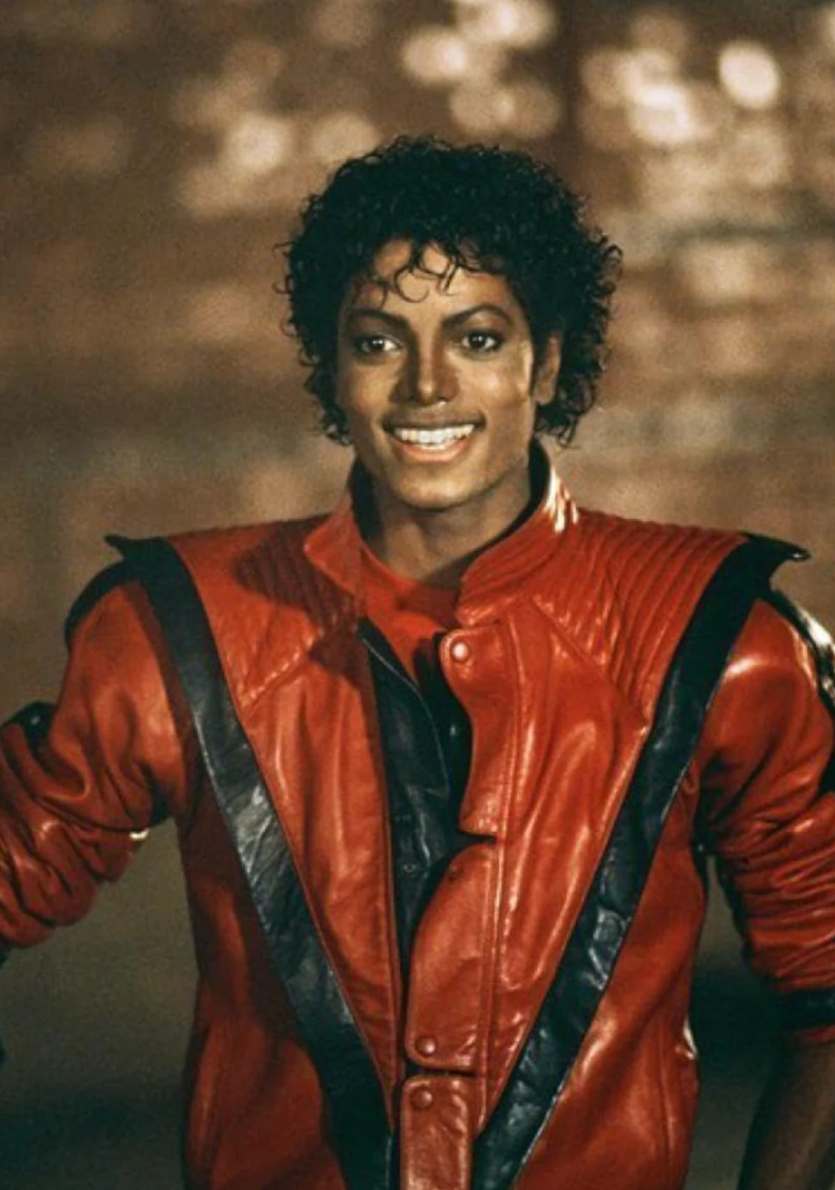 Kıvırcık siyah saçlara sahip, Thriller klibinden tanıdık kırmızı ve siyah detaylı deri ceketiyle genişçe gülümseyen Michael Jackson, bulanık kahverengi bir arka plan önünde neşeli bir ifadeyle duruyor.