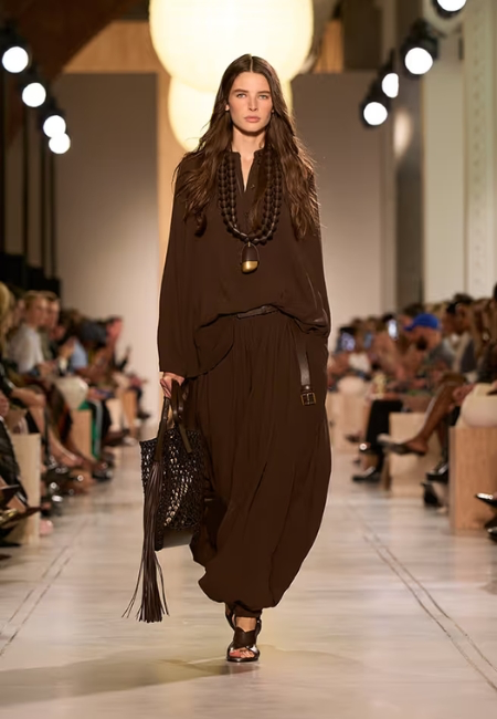 Michael Kors'un 2026 İlkbahar-Yaz koleksiyonundan bir model, kahverengi, bol kesim üst ve hafif kumaşlardan yapılmış hacimli balon pantolon takımıyla, uzun püsküllü örgü çantası ve boncuklu kolyesiyle podyumda yürüyerek bu sezonun dikkat çeken moda trendleri arasında yer alan akıcı silüetleri temsil ediyor.