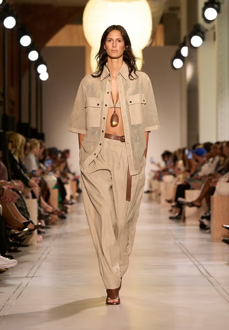 Podyumda, Michael Kors'un 2026 ilkbahar-yaz moda trendlerini yansıtan, bej tonlarında file detaylı gömlek, büstiyer ve hacimli balon pantolon giyen bir model, kahverengi kemeri, uzun sarkık kolyesi ve sandaletleriyle güçlü bir siluet sunuyor.