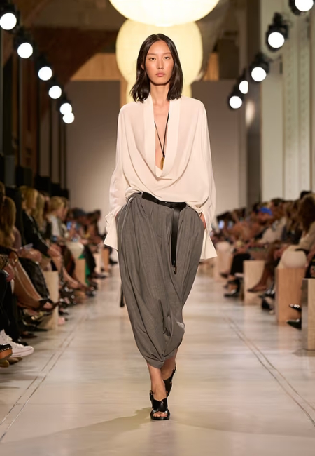 Michael Kors'un 2026 ilkbahar-yaz moda trendlerini yansıtan podyumda yürüyen bir model, derin V yakalı dökümlü krem bluz ve yeni bir siluet oluşturan hacimli gri balon pantolonlarla dikkat çekiyor.