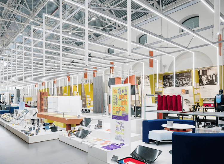 ADI Design Museum'daki Milano Design Week sergi alanından geniş bir görünüm; yüksek tavanı, beyaz lineer aydınlatmaları ve farklı yükseklikteki platformlarda sergilenen çağdaş tasarımları, arka planda sarı panellerdeki tarihi görseller ve İtalyanca afişlerle birleştirerek tasarımın geçmişi ve geleceği arasındaki diyaloğu gözler önüne seriyor.