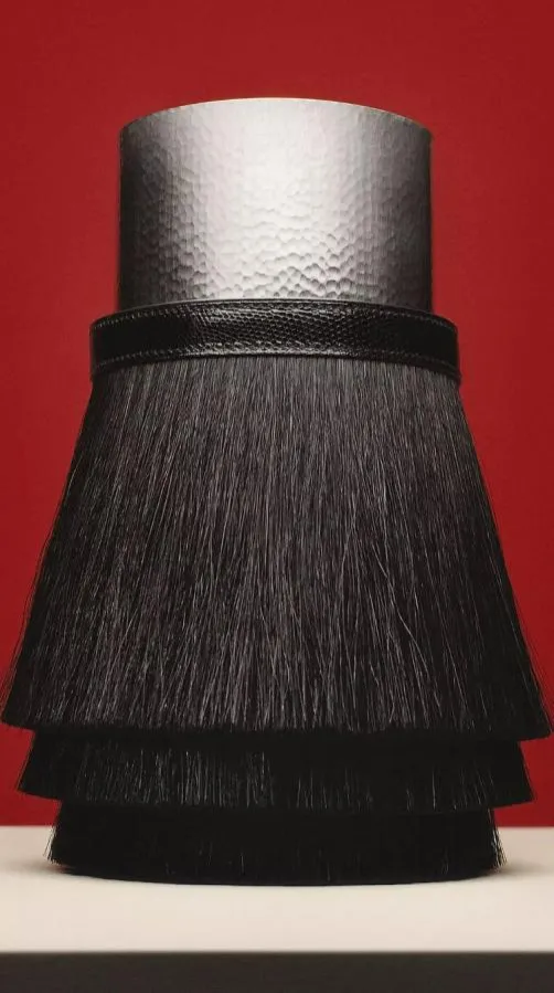 Milano Design Week kapsamında sergilenen, üst kısmı çekiçlenmiş gümüş renkli metal silindir, alt kısmında ise dar siyah deri bir bantla ayrılmış, katmanlı uzun siyah kıllardan oluşan, zanaatın ve lüks ev koleksiyonlarının inceliğini yansıtan estetik bir obje.