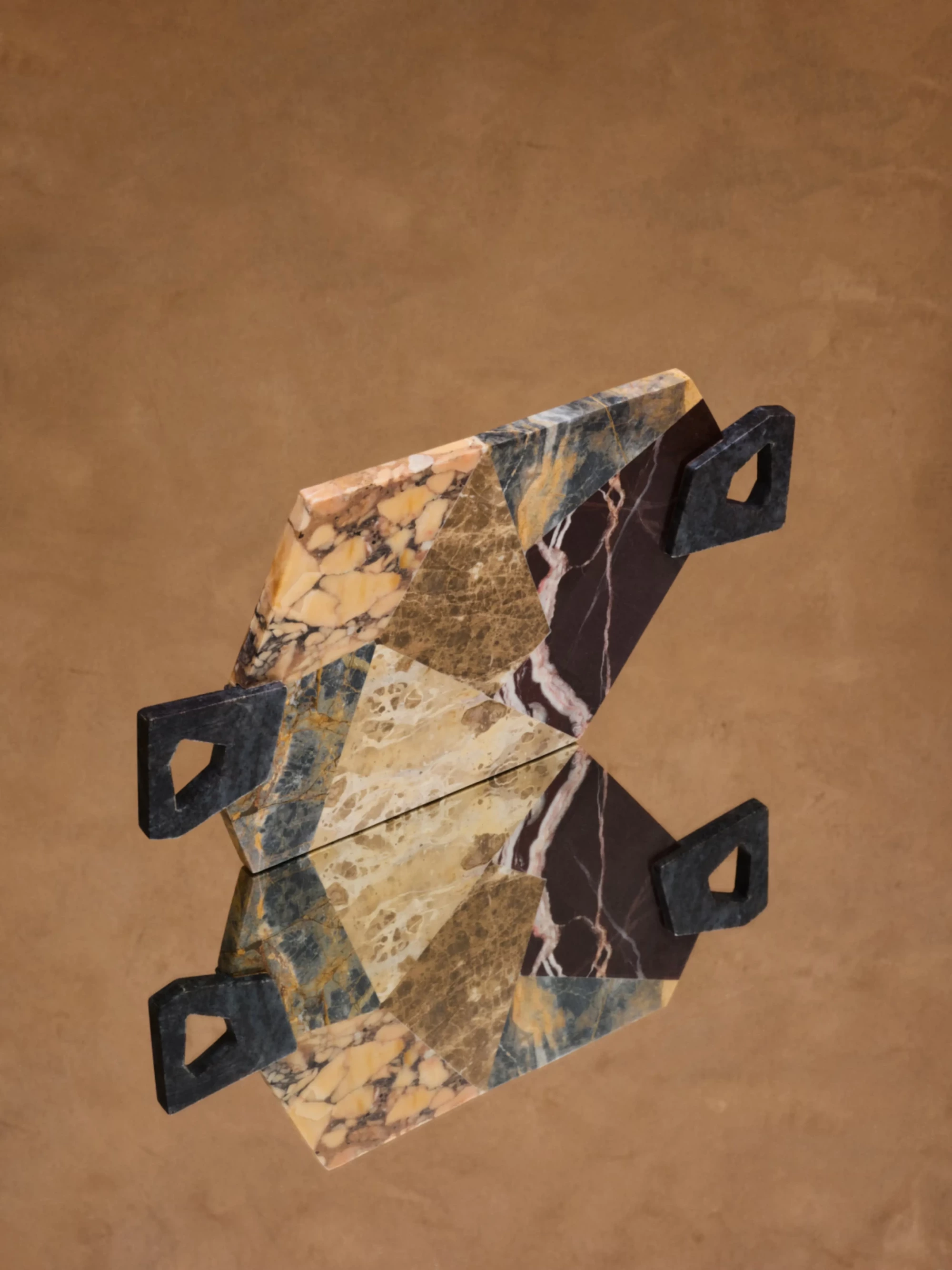 Milano Design Week 2026 kapsamında H&M Home için Kelly Wearstler tarafından tasarlanan, krem, kahverengi ve bordo tonlarındaki farklı mermer parçalarının geometrik birleşimiyle oluşan, siyah kulplu dekoratif bir obje, altında kendi yansımasıyla kahverengi bir zeminde lüks ve modern tasarım estetiğini sergiliyor.
