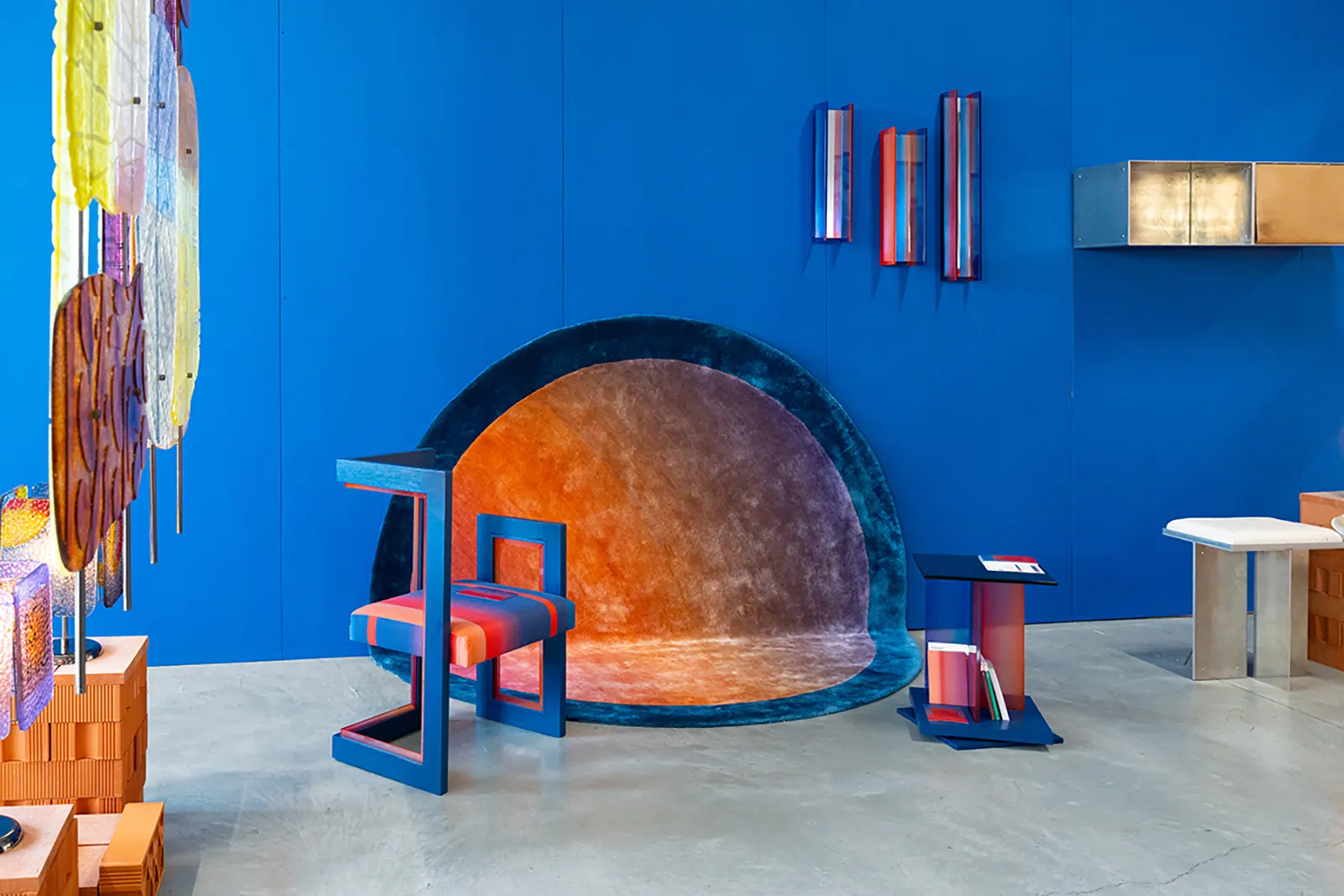 Milano Design Week 2026 kapsamındaki Isola Design Festival'inden bir kare, parlak mavi bir duvar önünde turuncu-mor geçişli yarım daire bir halı, çeşitli renklerdeki heykelsi aydınlatmalar ve cesur formlara sahip deneysel bir koltukla geleceğin yaşam formlarını sorgulayan modern tasarım denemelerini vurguluyor.