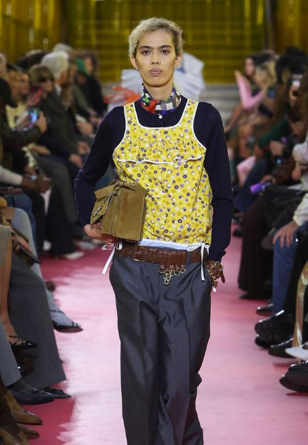 Model, 2026 ilkbahar-yaz moda trendleri kapsamında Miu Miu koleksiyonundan; lacivert uzun kollu triko üzerine giydiği canlı sarı çiçek desenli fırfırlı askılı bluzu, koyu gri pantolonu, kahverengi deri kemeri ve süet çantasıyla podyumda sergiliyor. Bu kombin, sezonun renk blokları ve günlük stile adapte edilen enerjik siluetlerini öne çıkarıyor.