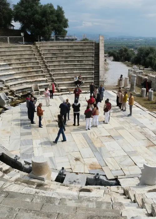 Bir antikkentteki basamaklı taş oturma yerleri ve mermer zeminli orkestra alanında, bir grup ziyaretçi tarihi kalıntıları dikkatle incelerken, arka planda zeytin ağaçları ve uzaklardaki yeşil vadi bulutlu bir gökyüzü altında uzanıyor.