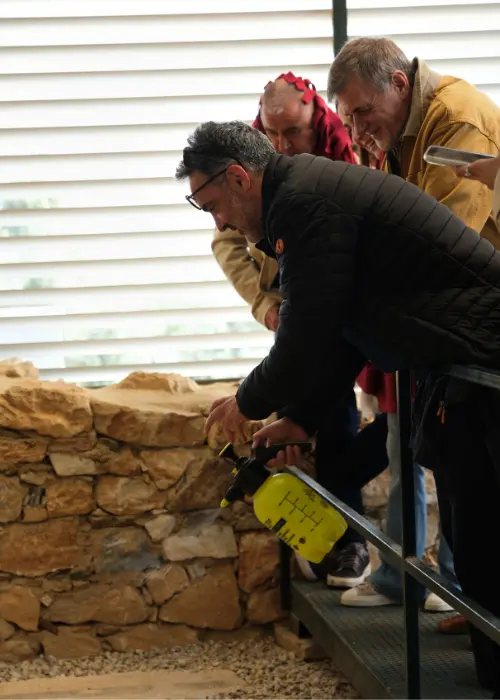 Siyah montlu ve gözlüklü bir adam, sarı bir püskürtücü kullanarak antik taş bir duvarı dikkatle temizlerken, arkasındaki iki kişi bu özenli anı izliyor ve bu görüntü bir antikkentin korunma veya araştırma sürecindeki hassas çalışmaları yansıtıyor.