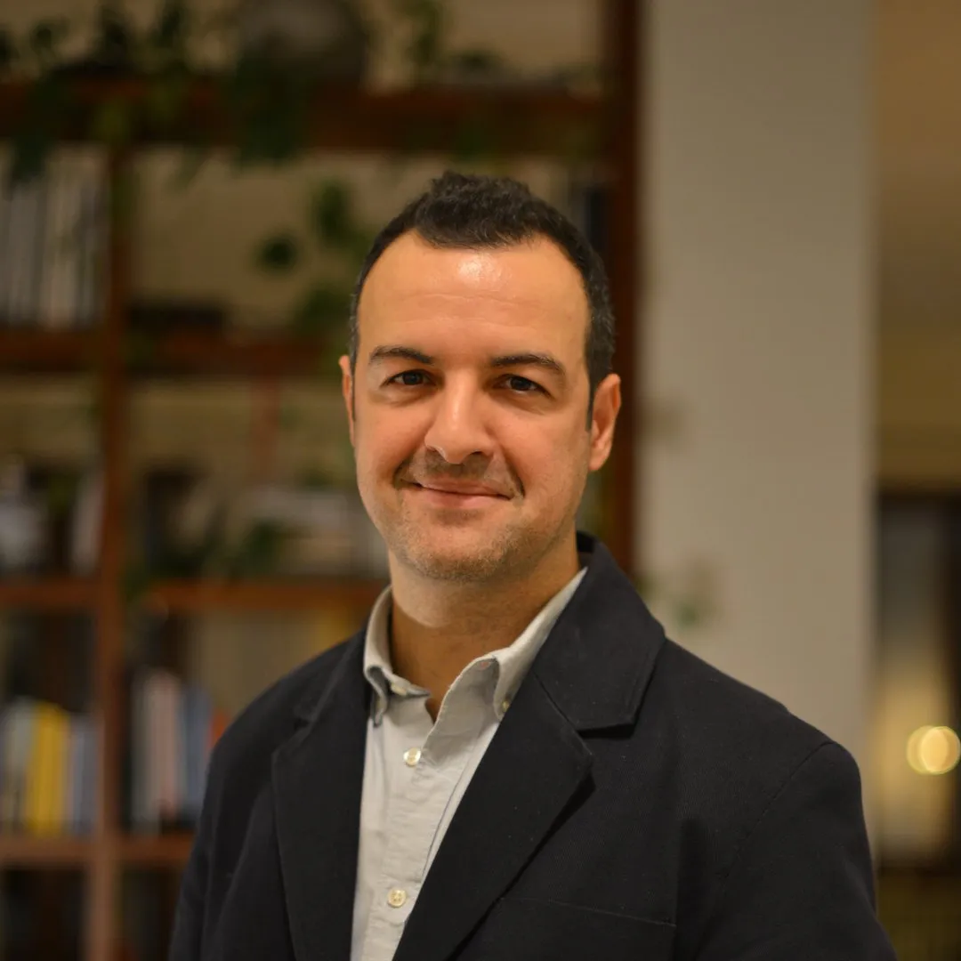 Gazeteci Murat Pazarbaşı, Milano Design Week yorumlarını paylaştığı yazısında, hafif gri gömleği ve koyu lacivert ceketiyle kameraya gülümseyerek bakarken, arka planda bulanık bir kitaplık ve yeşil bitkiler görülüyor.