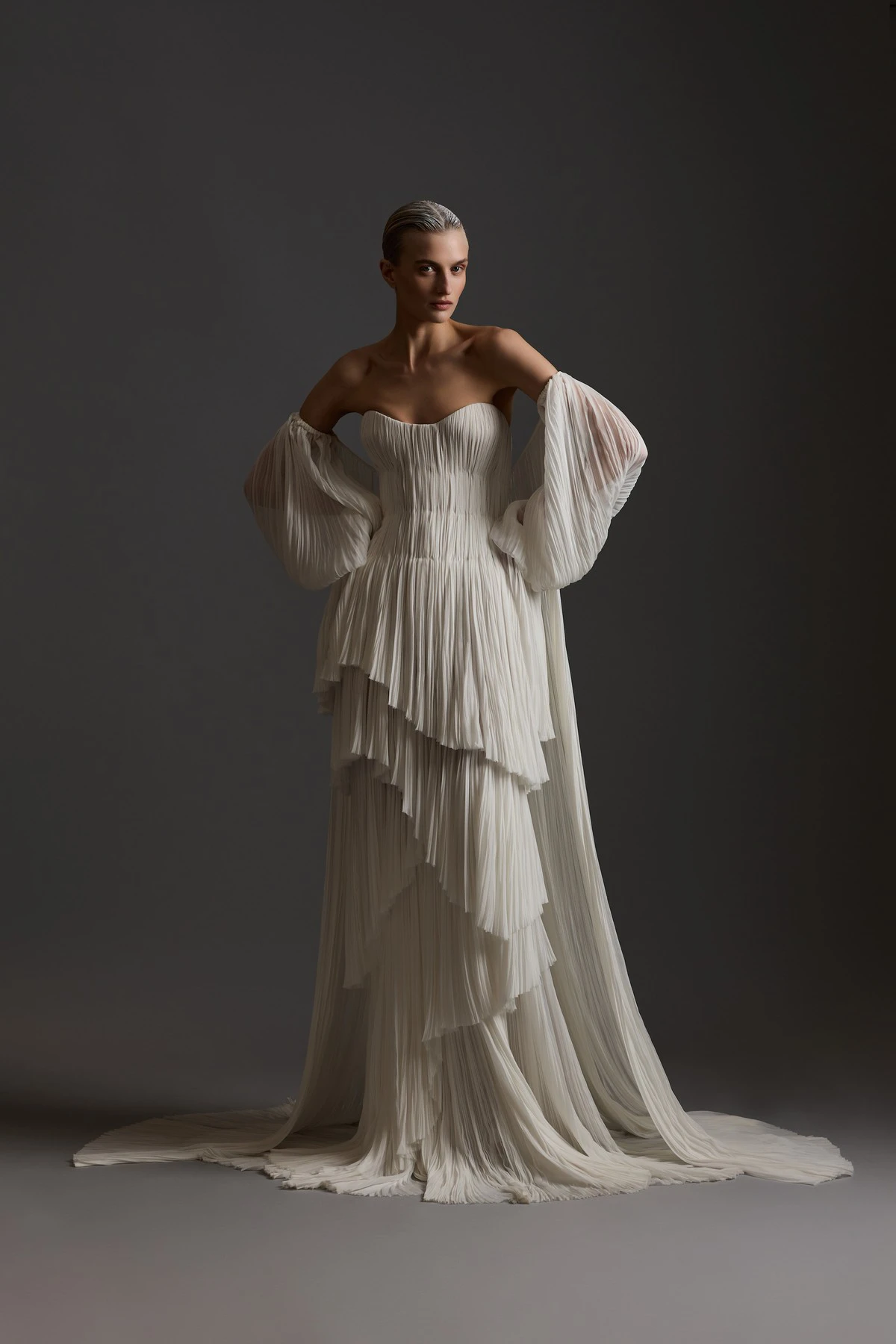New York Bridal Moda Haftası'nın 2027 İlkbahar gelinlik koleksiyonları içinde öne çıkan bir trendi temsil eden bu görselde, fildişi rengi, kat kat pileli, straplez bir Elie Saab gelinliği giyen model, hacimli pelerin benzeri kolları ve uzun, yere uzanan etekleriyle zarif bir duruş sergiliyor.