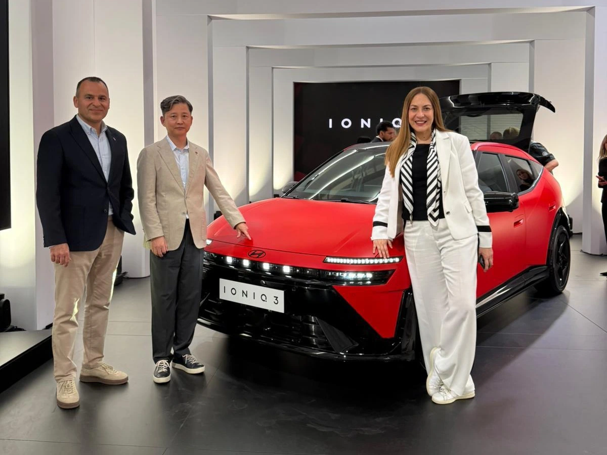 Milano'da gerçekleşen Hyundai IONIQ 3 lansmanı etkinliğinde, şık beyaz takımıyla Özlem Gusar ve iki yönetici, kırmızı renkli yeni nesil elektrikli otomobilin önünde poz veriyor, ortadaki yönetici aracın kaputunu işaret ediyor.