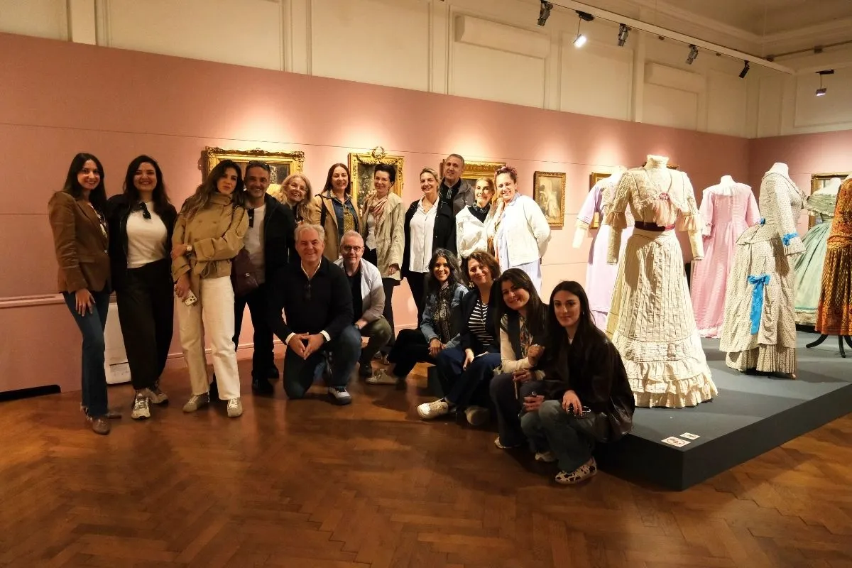 OGGUSTOGETHER etkinliği kapsamında Arkas Sanat'ın bir mekanında çekilen bu grup fotoğrafında, OGGUSTO ekibi ve davetli konuklar pembe duvarlar önünde, çerçeveli tablolar ve mankenler üzerinde sergilenen zarif dönem kıyafetleri arasında neşeli bir şekilde poz veriyorlar.
