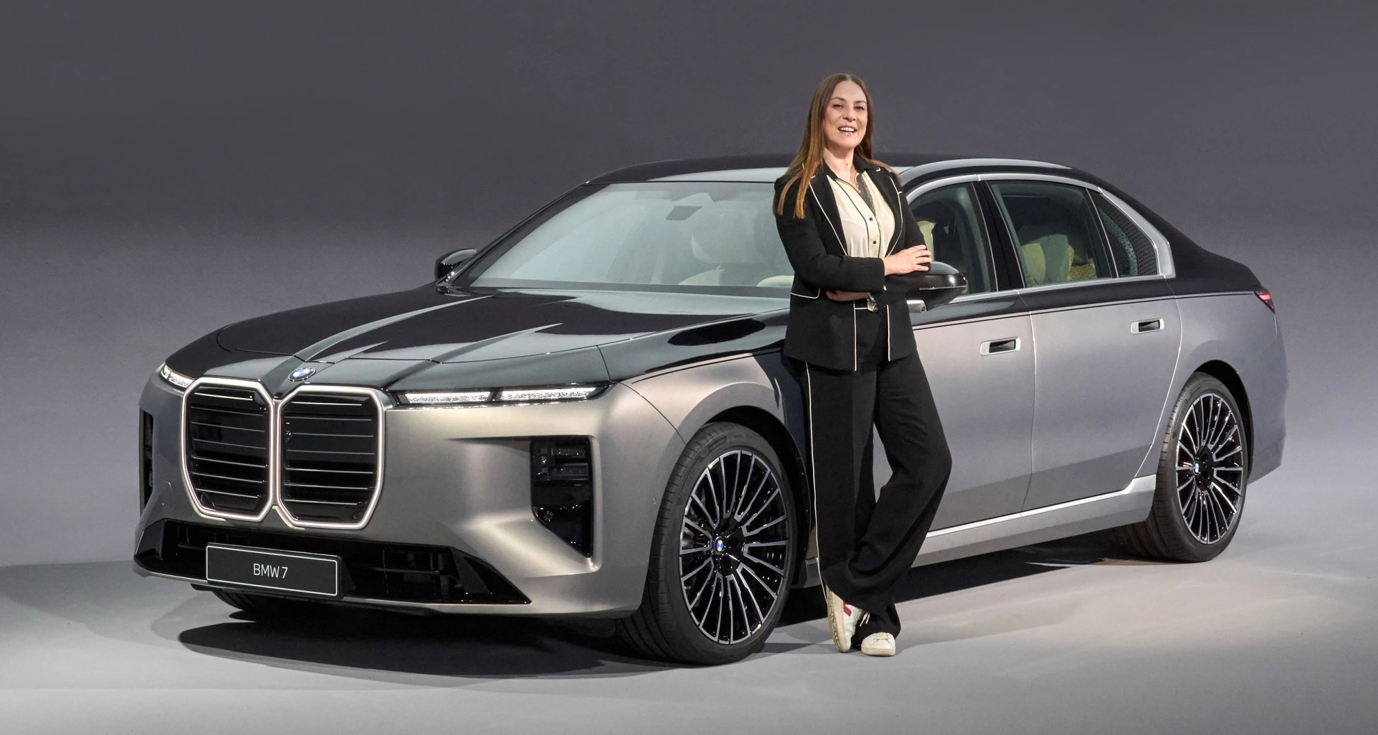 Özlem Gusar, yeni BMW 7 Serisi i7 modeliyle olan deneyimini yansıtan bu fotoğrafta, koyu gri ve açık gri tonlarda iki renkli tasarımıyla dikkat çeken lüks otomobilin yanında kollarını kavuşturmuş ve gülümseyerek poz veriyor.