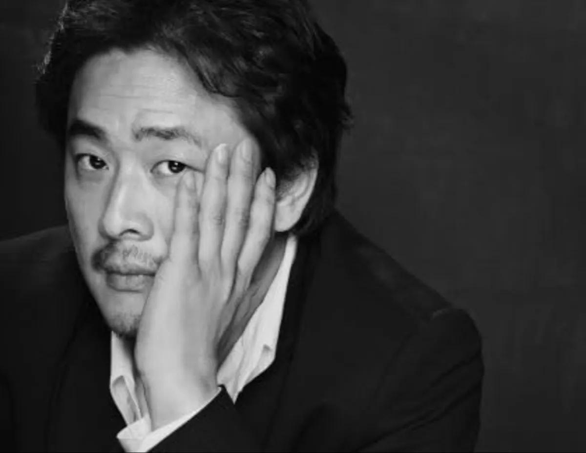 Siyah beyaz Park Chan-wook portresi, Güney Koreli film yönetmeni yüzünün bir kısmını sol eliyle kaplamış, düşünceli bir ifadeyle doğrudan kameraya bakarken, üzerinde açık yakalı beyaz gömlek ve koyu renk bir ceket bulunuyor.