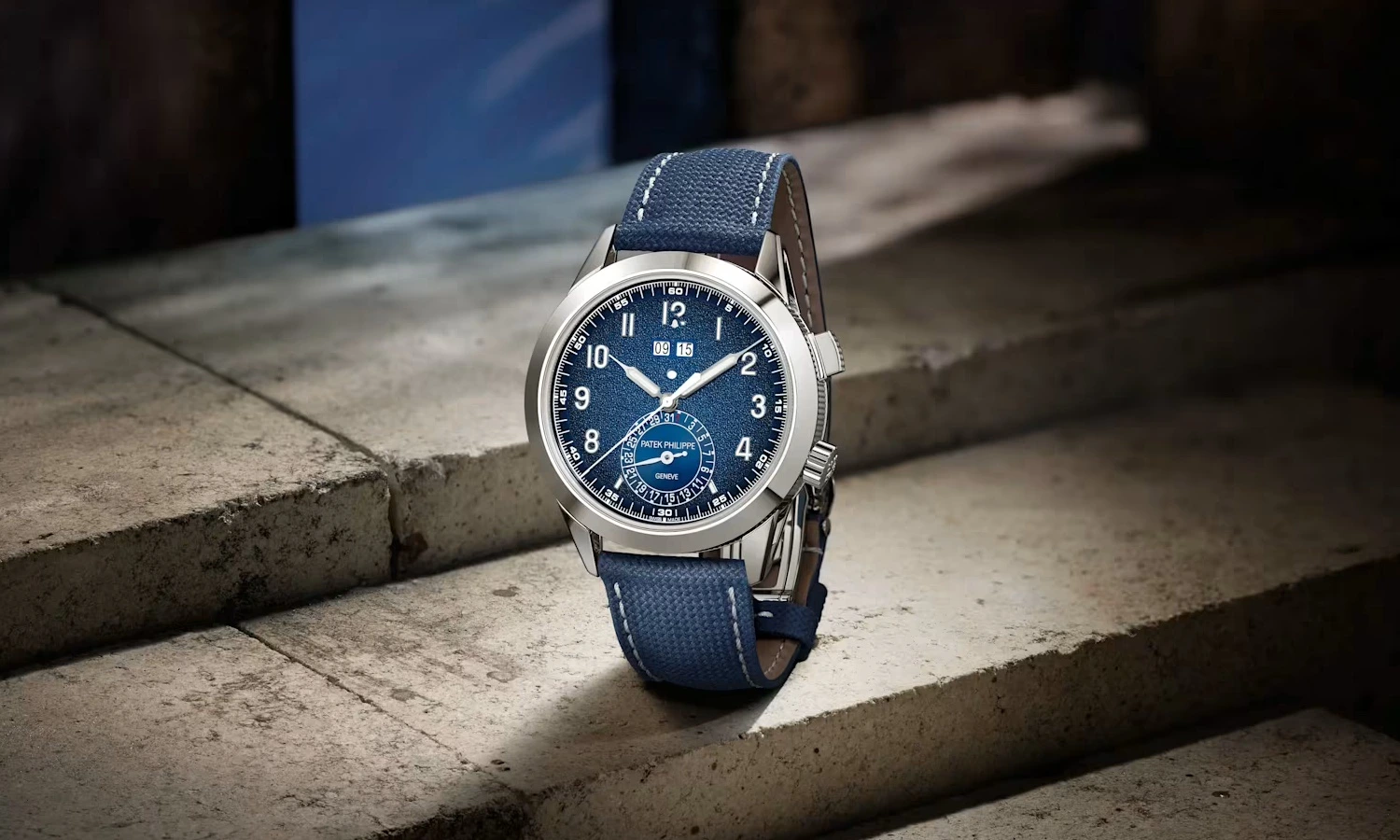 Cilalı gümüş renkli kasaya, derin dokulu mavi kadrana ve uyumlu mavi kanvas kayışa sahip, brüt beton basamaklar üzerinde zarifçe sergilenen bir Patek Philippe 5322G alarm saatinin yakın çekim görüntüsü.