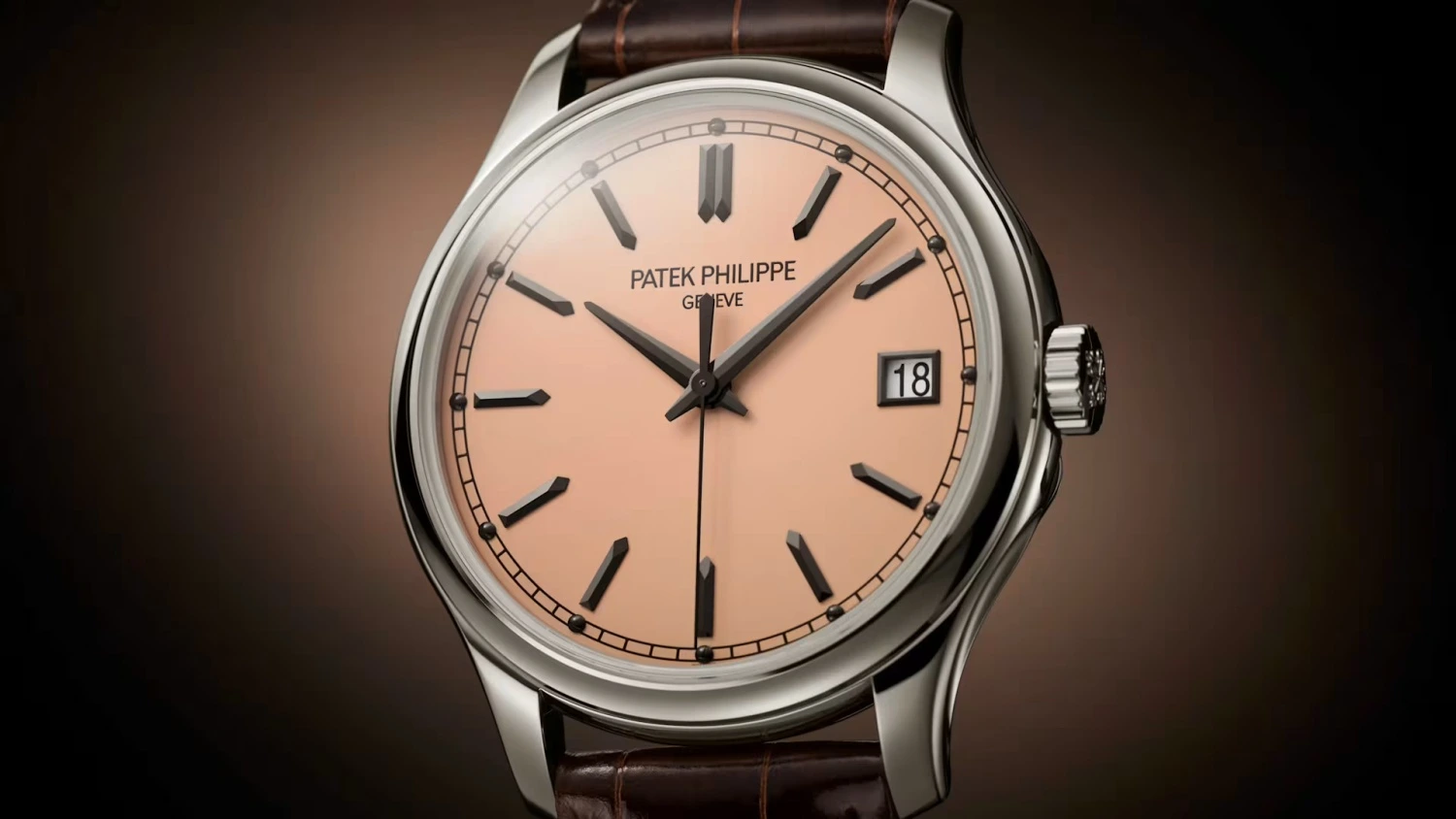 Patek Philippe Calatrava 5227G model kol saati, somon rengi kadranı, zarif beyaz altın kasası ve koyu kahverengi timsah derisi kayışıyla, saat 3 konumundaki 18 rakamını gösteren tarih penceresiyle dikkat çekiyor ve Watches and Wonders etkinliği kapsamında sergilenebilecek modern bir klasik.