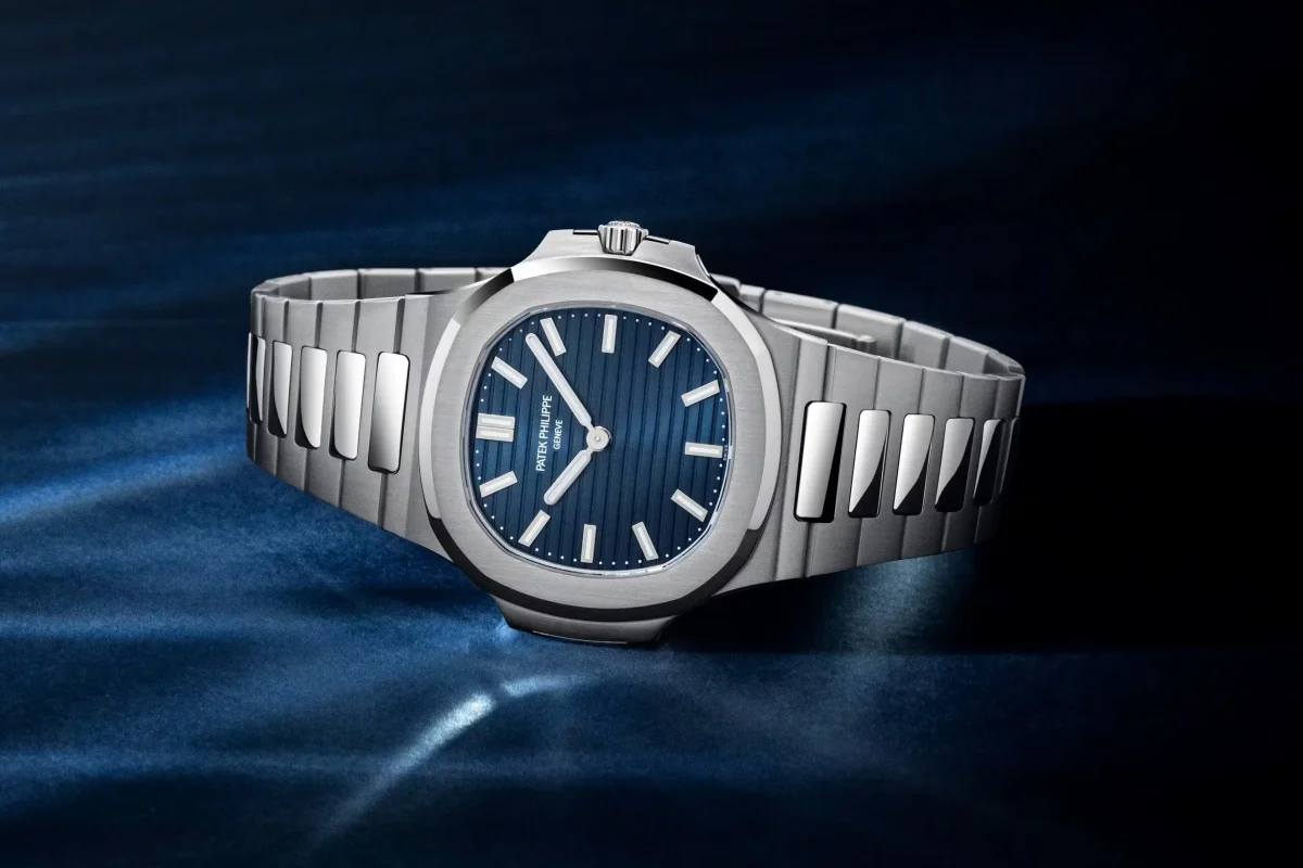 Koyu mavi yatay çizgili kadranı ve belirgin sekizgenimsi kasasıyla ikonik Patek Philippe Nautilus modelinin, 50. yıl dönümünü kutlayan gümüş renkli metal bilezikli zarif bir versiyonu, ışık yansımalarının olduğu koyu mavi tonlarındaki bir zemin üzerinde dikkat çekici şekilde duruyor.