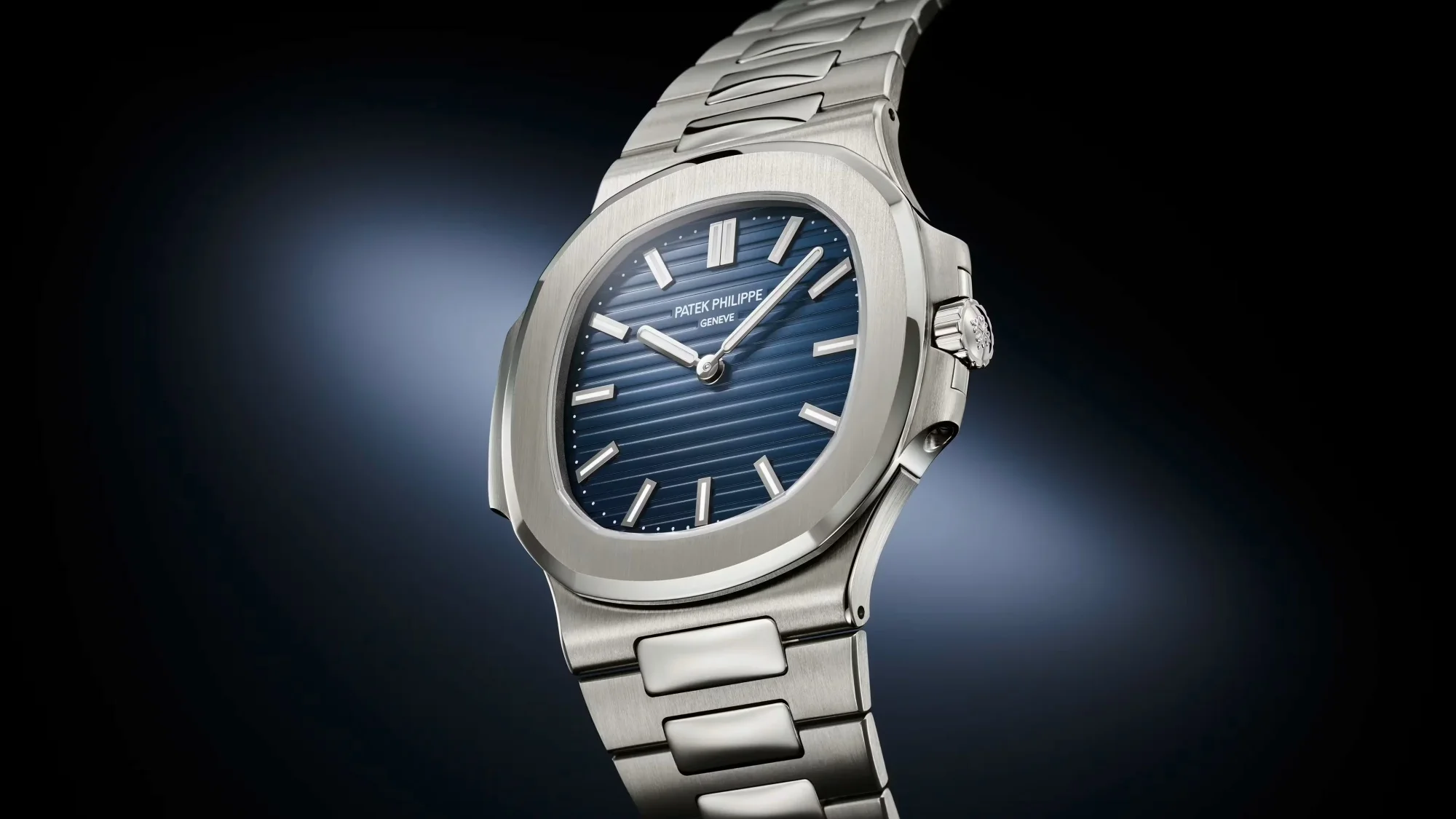 Patek Philippe Nautilus 50. yıl özel saati, yatay çizgili lacivert kadranı, sekizgen çelik kasası ve entegre çelik bileziğiyle güçlü bir ışık altında karanlık fonda göz alıcı biçimde sergileniyor.