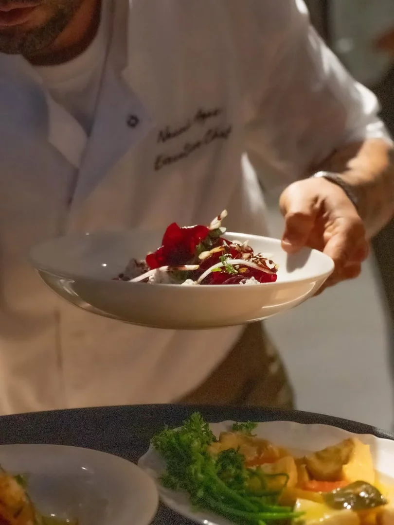 Beyaz şef ceketli bir aşçı, üzerinde ince dilimlenmiş pancar, peynir ve badem dilimleri bulunan renkli bir tabağı ustaca sunarken, ceketinde Naomi Ayaz Executive Chef yazısı seçiliyor ve bu an Peperino Bodrum Çarşı deneyimini yansıtıyor.