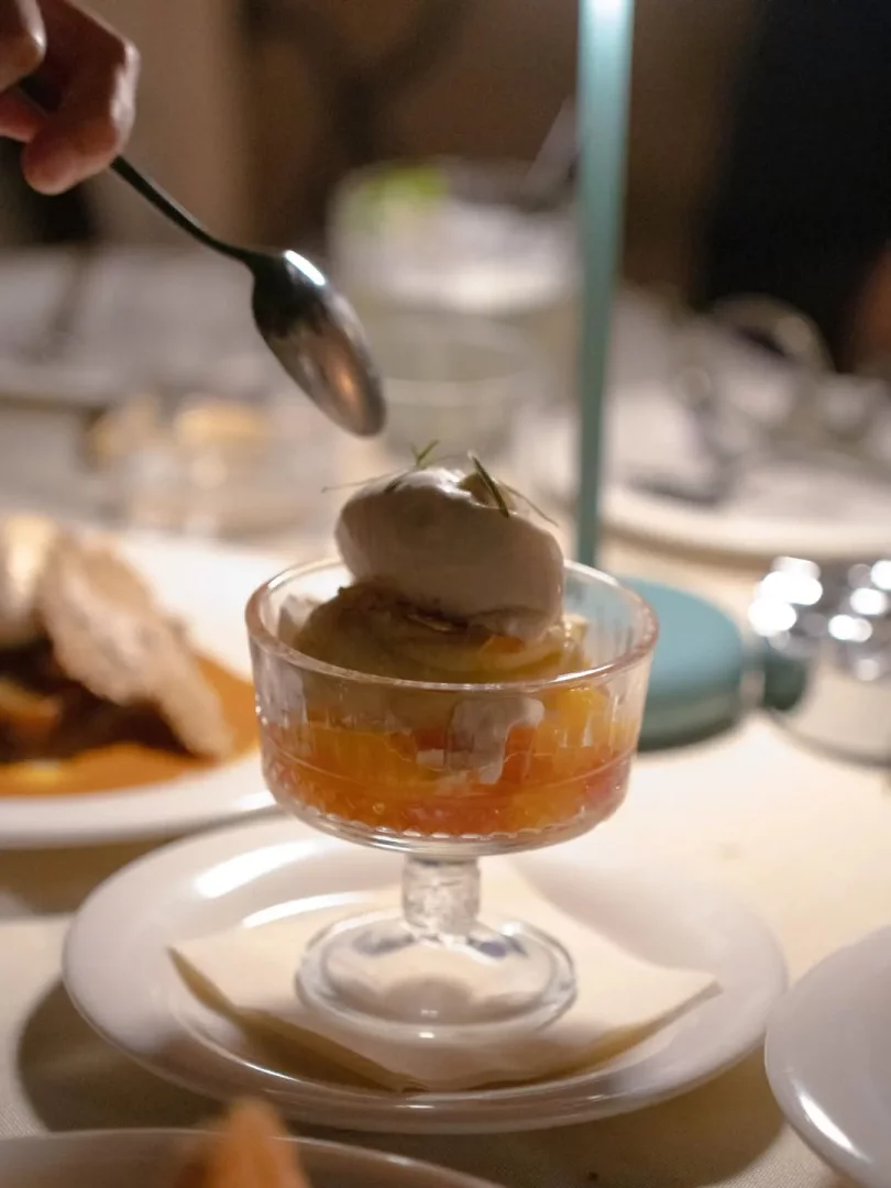 Bodrum Çarşı'daki Peperin restoranında akşam yemeği sonrası tatlı anı: masada duran şık cam kadehte, alt katmanı parlak turuncu renkli meyve taneleri, üzerinde açık renkli bir dondurma topu veya krema ve küçük bir yeşil dal ile süslenmiş zarif bir tatlının, gümüş bir kaşıkla servis edilmeye hazırlandığı yakın çekim bir görüntü.