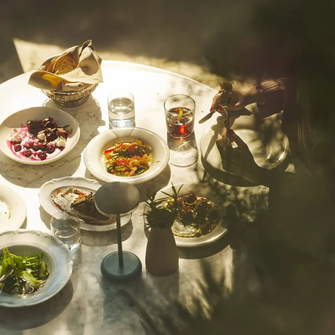 Bir Bodrum çarşı restoranında güneşli bir masada, kırmızı meyveli tatlı, balık, çeşitli salatalar, içecekler ve ekmek sepetiyle zengin bir öğle yemeği veya akşam yemeği menüsü, bir kişinin yemek yemeye hazırlanırken elleriyle çatal bıçak tuttuğu keyifli bir an.