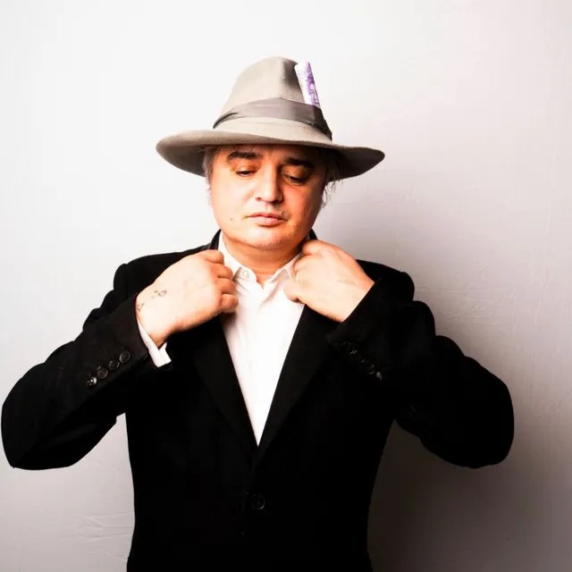 Açık gri fötr şapkasına mor renkli bir not veya bilet sıkıştırmış olan Peter Doherty, griye çalan saçları ve siyah ceketiyle beyaz gömleğinin yakasını iki eliyle düzeltirken aşağıya bakıyor.
