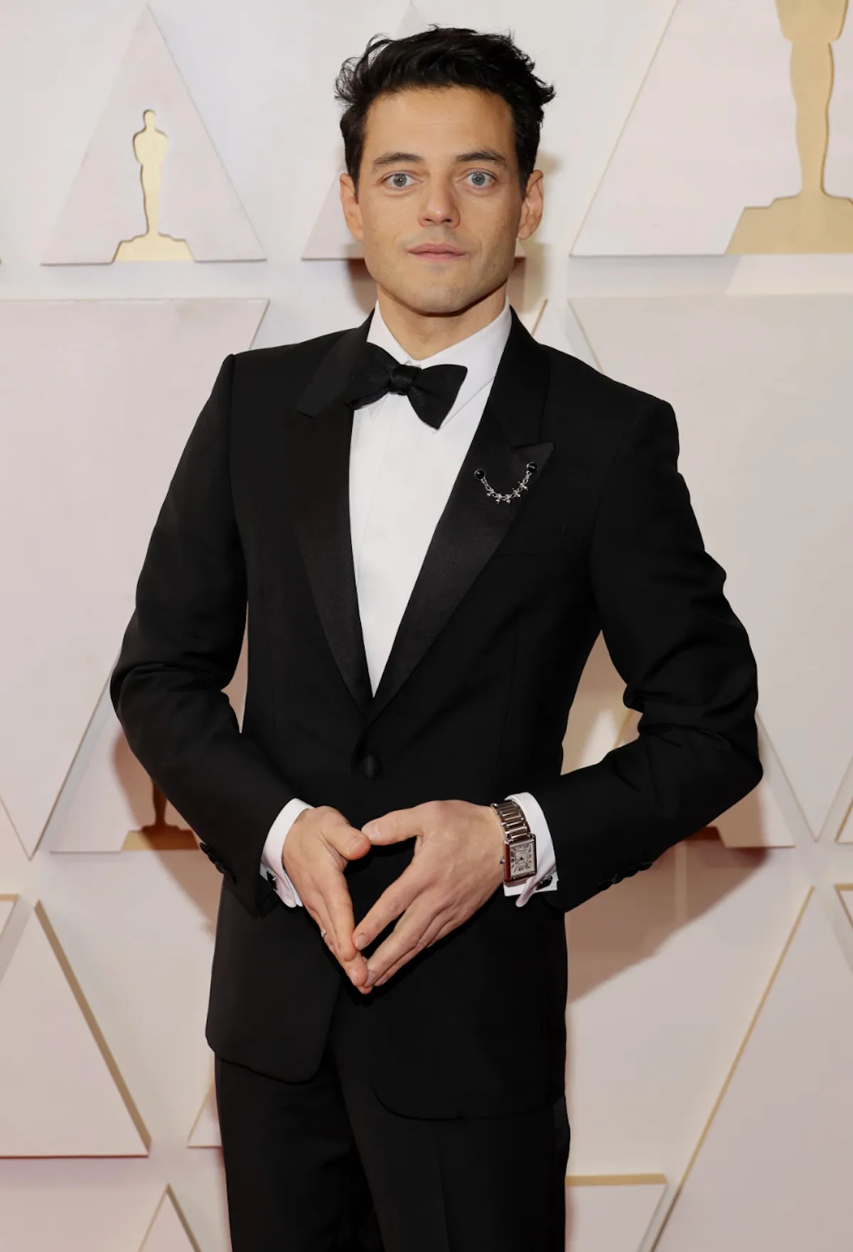 Rami Malek, 2022 Oscars kırmızı halısında siyah smokini, beyaz gömleği ve papyonuyla, sol yakasında zincir detaylı bir broş ve sol bileğinde dikdörtgen gümüş bir saatle, ellerini önünde kalp şeklini andıracak şekilde birleştirmiş, arka planda Oscar heykeli siluetleri varken objektife nötr bir ifadeyle bakıyor.