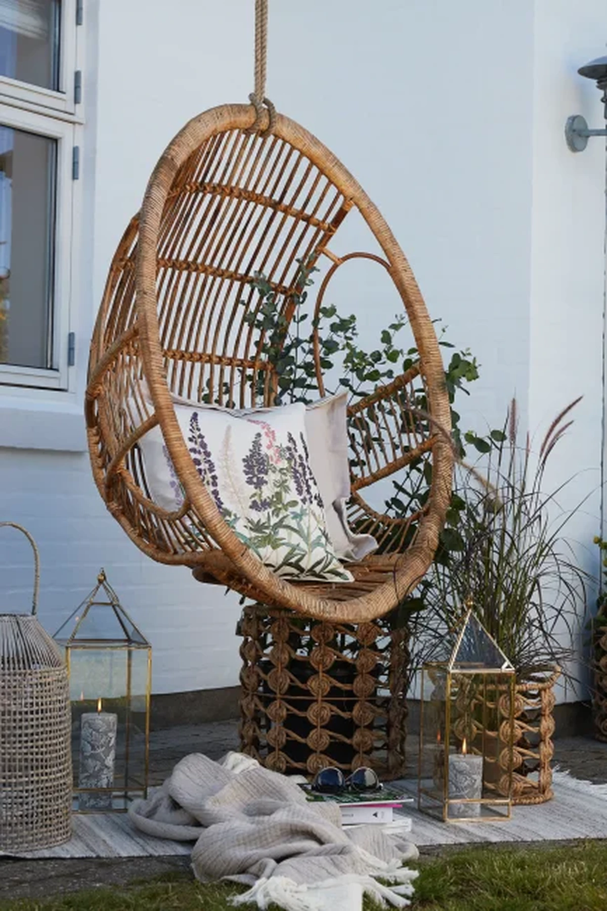 Dış mekanın huzurlu atmosferinde, beyaz bir duvarın önünde asılı duran, çiçek desenli bir yastıkla süslenmiş konforlu bir rattan salıncak, çevresindeki şık fenerler ve yeşil bitki tasarımlarıyla doğal ve davetkar bir köşe oluşturuyor.