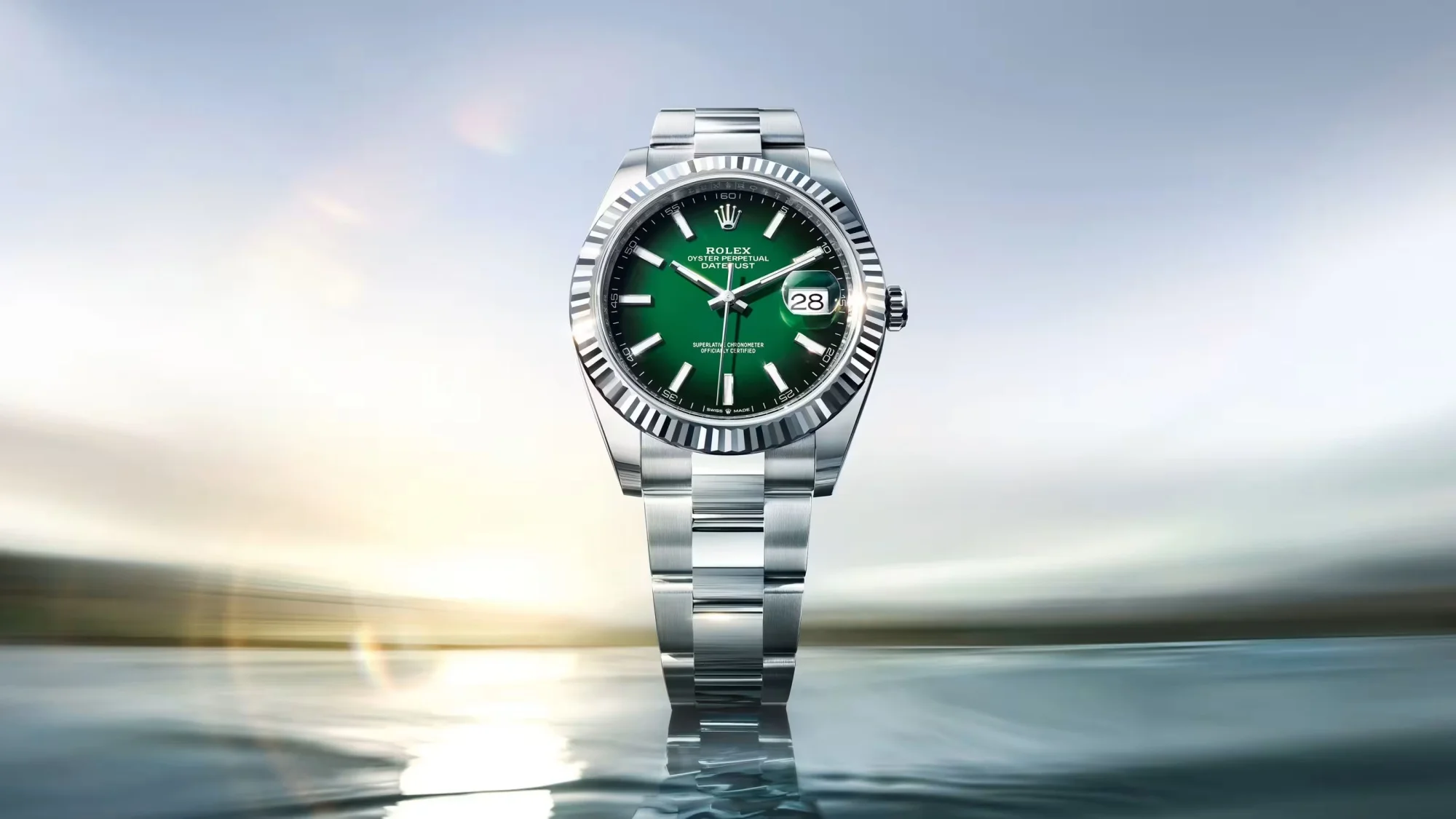 Parlak, yansıtıcı bir zeminde, aydınlık ve geniş bir arka plan önünde, canlı yeşil kadranı, klasik yivli bezeli ve zarif metal bileziğiyle dikkat çeken bir Rolex Datejust saati, tarih penceresinde 28'i gösterirken şık bir şekilde sergileniyor.