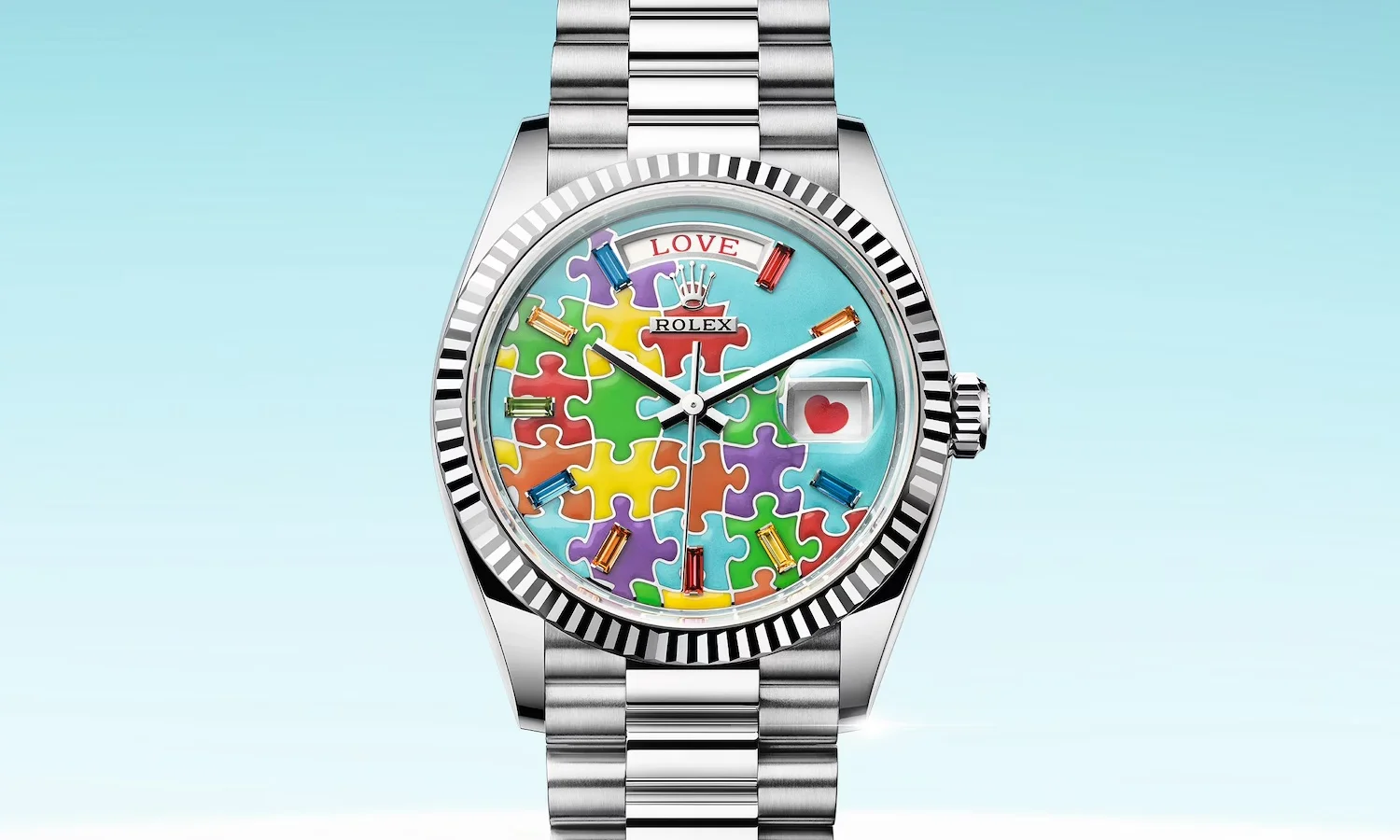 Açık mavi bir arka plan üzerinde, benzersiz tasarıma sahip beyaz altın Rolex Day-Date kol saati, çok renkli puzzle parçaları ve farklı renklerde baget taşlarla süslenmiş mavi kadranı, 3 konumunda kırmızı kalp detayı ve 12 konumunda LOVE yazısıyla dikkat çekiyor.