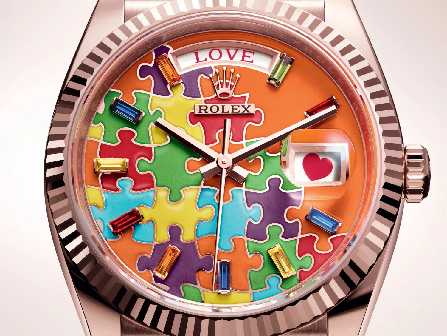 Yakın planda, pembe altın kasalı Rolex Day-Date saatin çok renkli yapboz desenli turuncu kadranı, LOVE yazısı, 12 konumundaki Rolex tacı, parlak renkli baget kesim değerli taşlardan oluşan saat imleri ve 3 konumundaki kırmızı kalp simgesi gösteren tarih penceresiyle detaylı olarak sunuluyor.