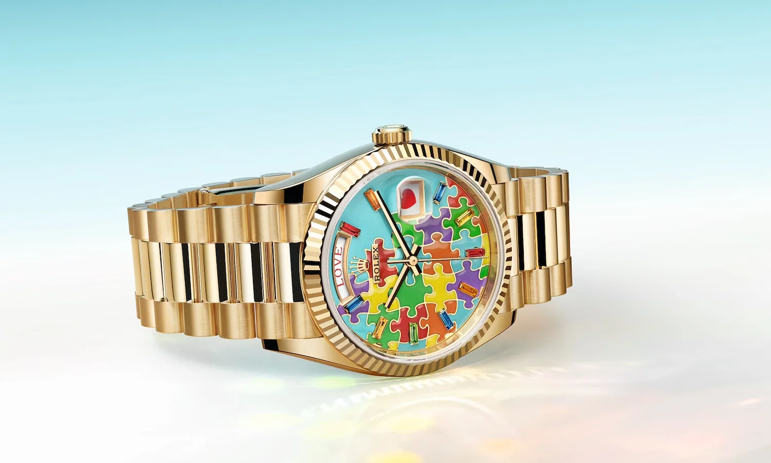 Sarı altın rengi Rolex Day-Date saatin, açık mavi zeminli, canlı renkli puzzle parçaları ve çok renkli taşlarla süslü kadranında saat 12 yönünde kırmızı kalp, saat 9 yönünde ise LOVE yazısı dikkat çekiyor, tırtıllı bezel tasarımıyla lüks bir görünüm sunuyor.