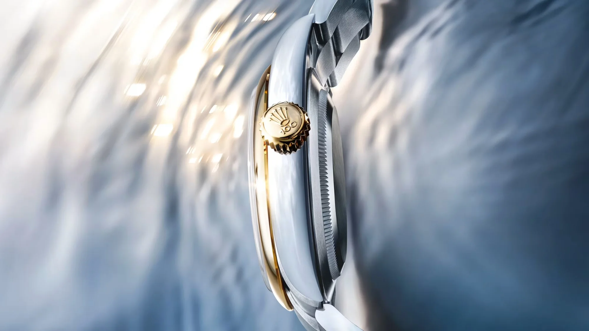 Parıltılı ışıklar yansıtan, hafifçe bulanık gri-mavi bir zeminde, Rolex Oyster Perpetual serisine ait, altın bezelli ve tepe kısmında Rolex logosu ile birlikte 100 sayısı kabartmalı olarak işlenmiş lüks bir kol saatinin yandan yakın çekimi.
